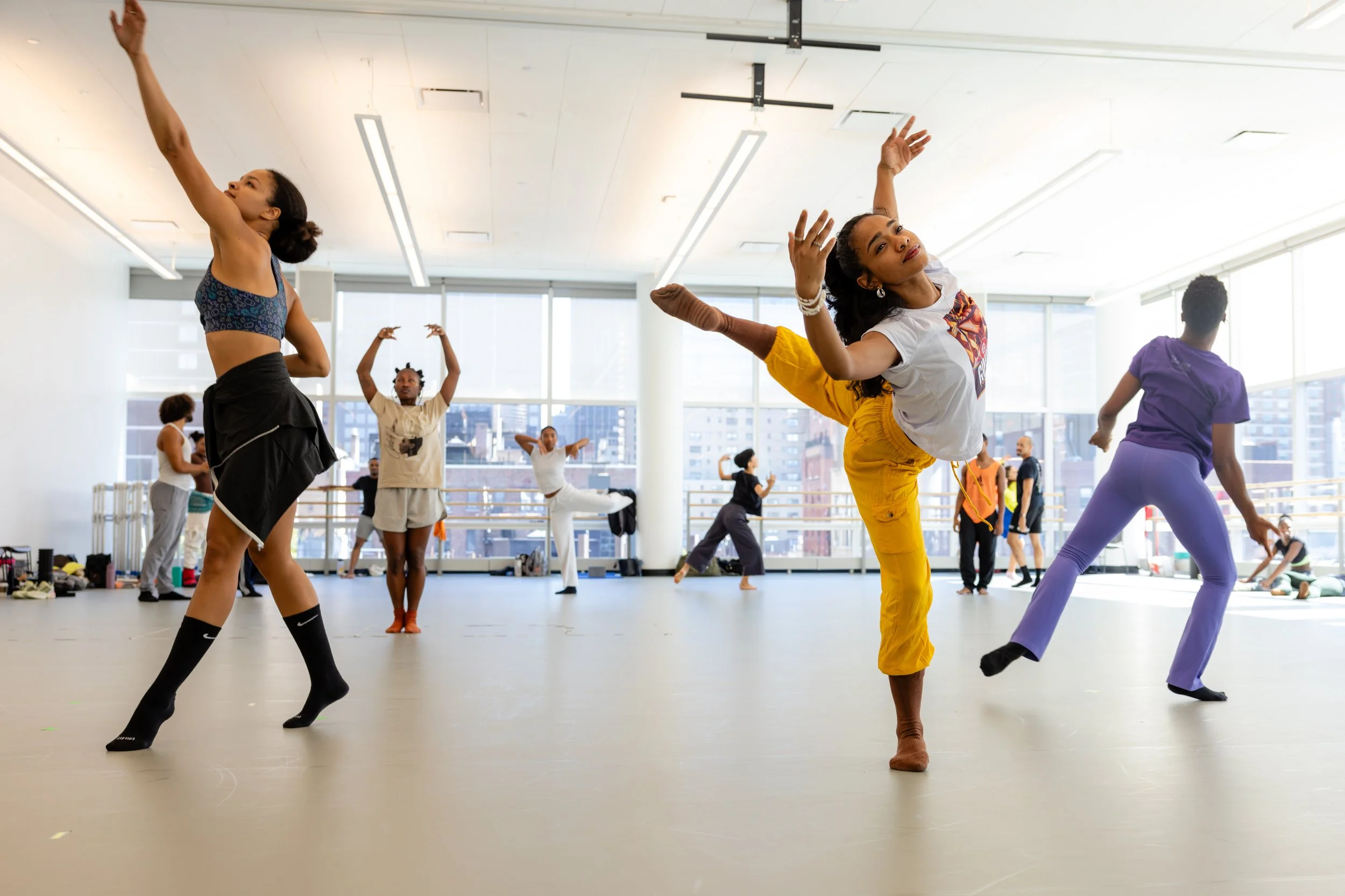 People practicing dance or fitness in a brightly lit studio with large windows, some lifting their arms and balancing on one leg.