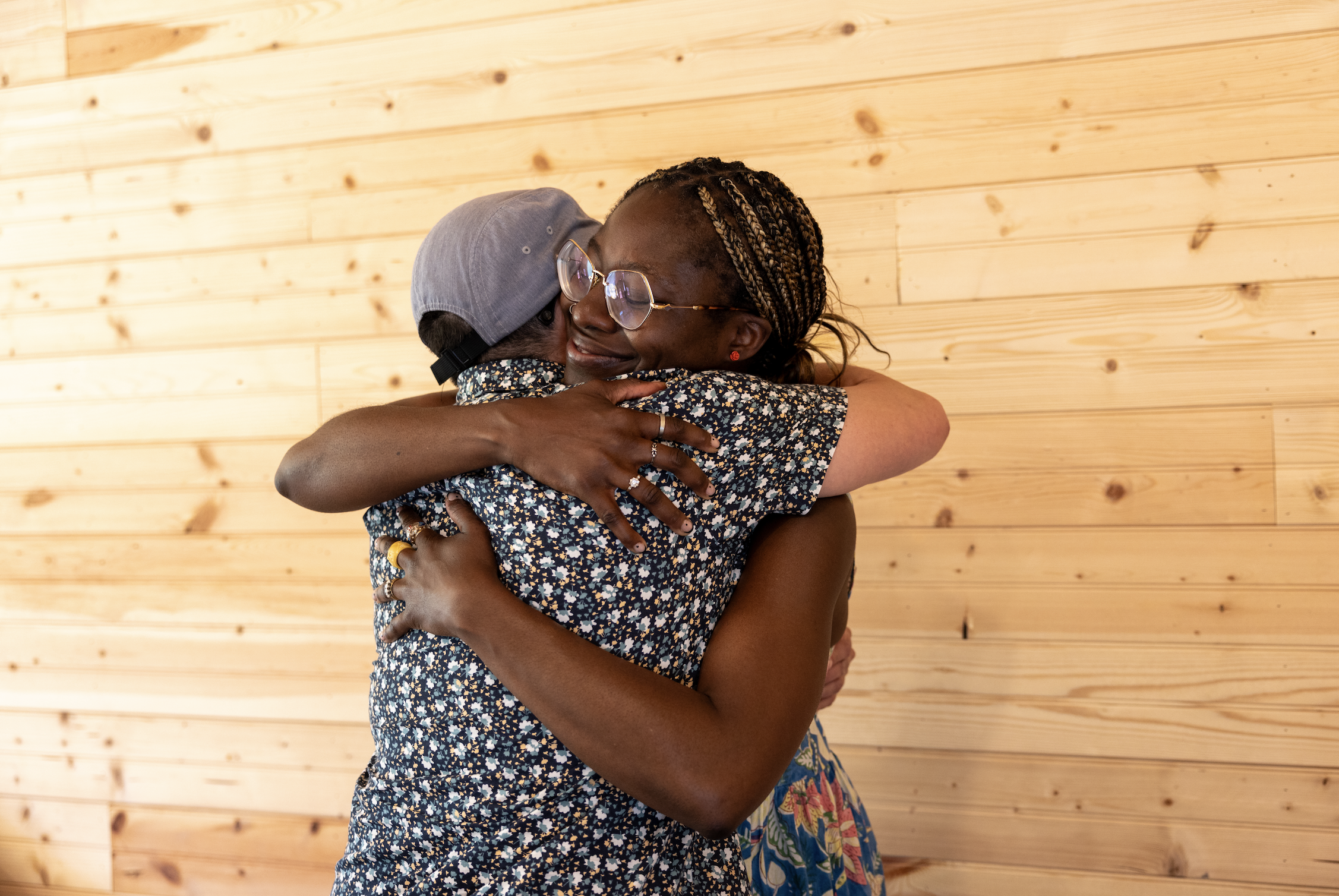 Two women hugging each other with smiles, standing in front of a wooden wall.