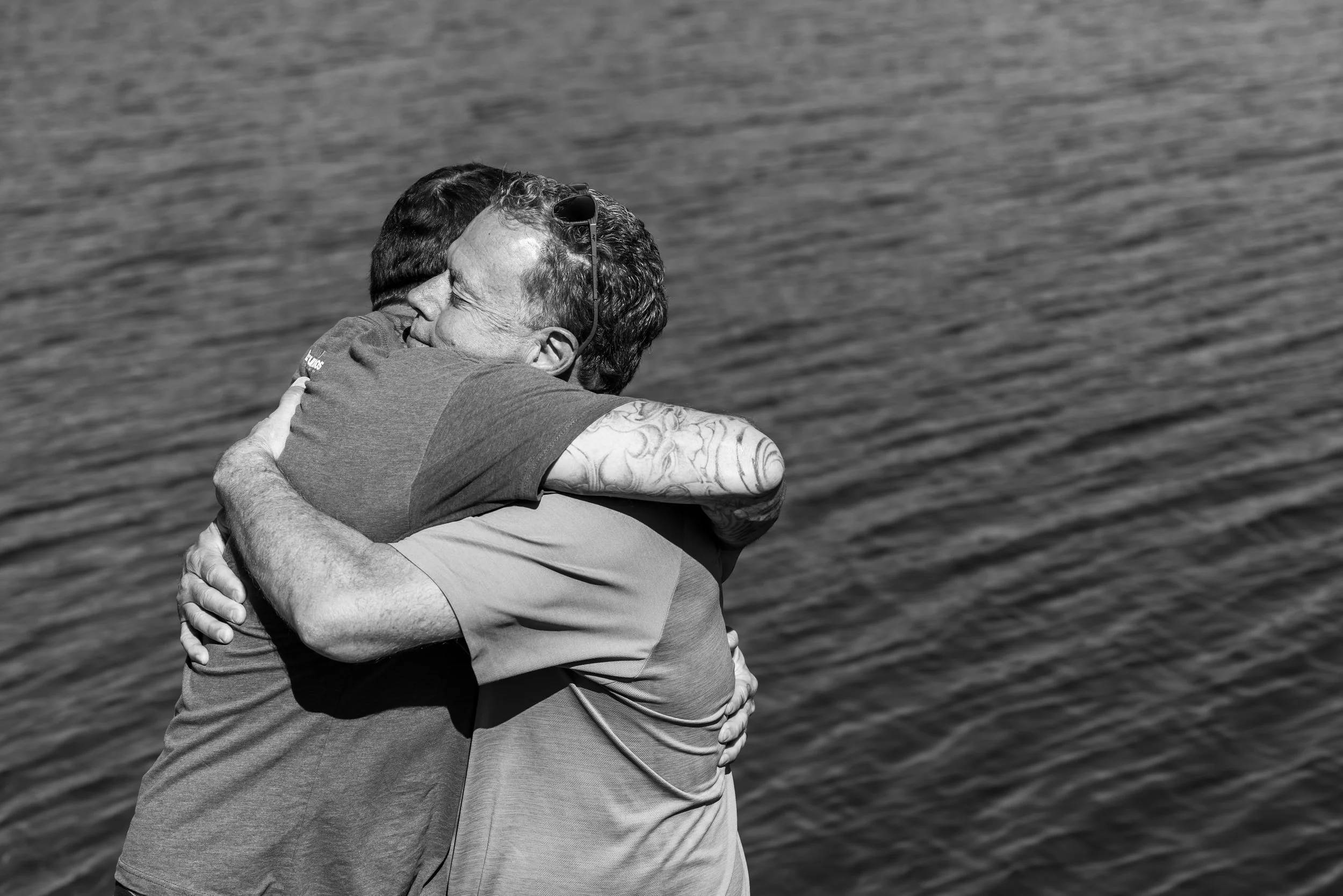 Two men embracing each other and smiling, standing near a large body of water.