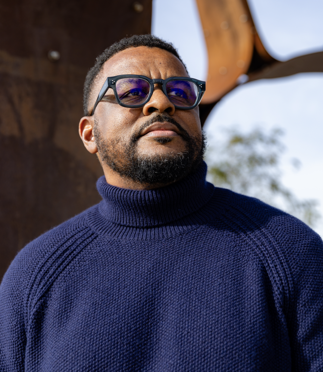 A man with dark hair, beard, and glasses wearing a blue turtleneck sweater, standing outdoors against a rusty metal structure with a clear sky in the background.