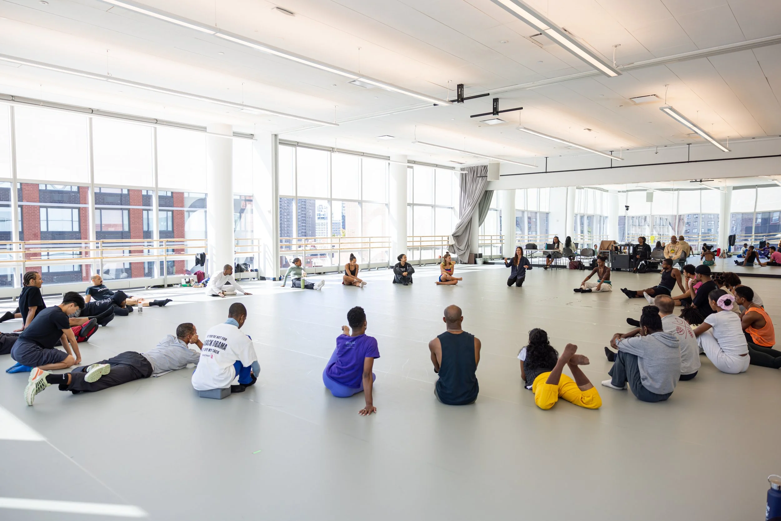A large group of people participating in a dance or exercise class inside a spacious, bright studio with floor-to-ceiling windows and a cityscape view.