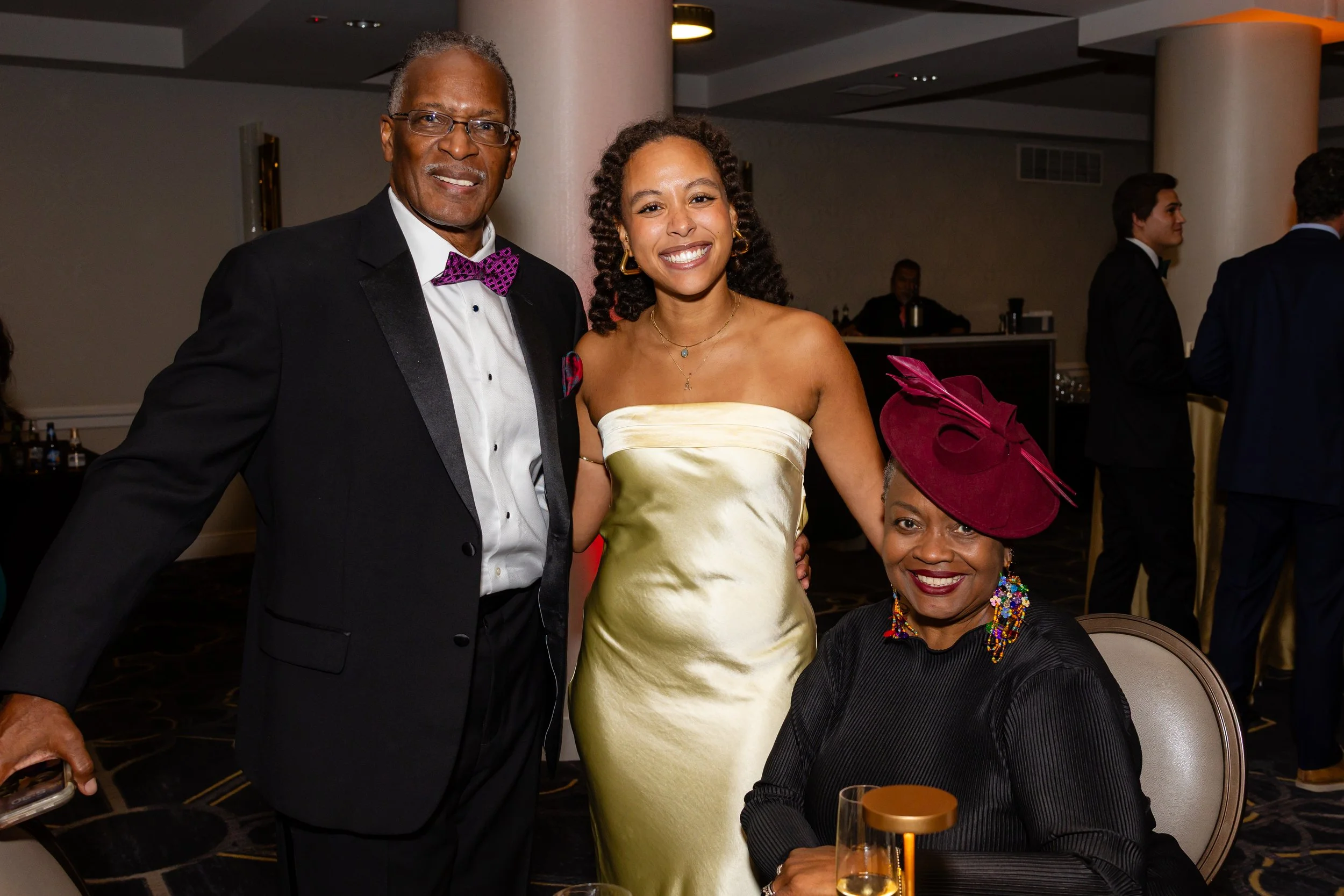 Four people at a formal event, three women and one man. The man is wearing a tuxedo with a purple bow tie, and the women are in elegant dresses, one holding a bouquet of flowers. They are smiling and posing for the photo. Other guests are visible in 