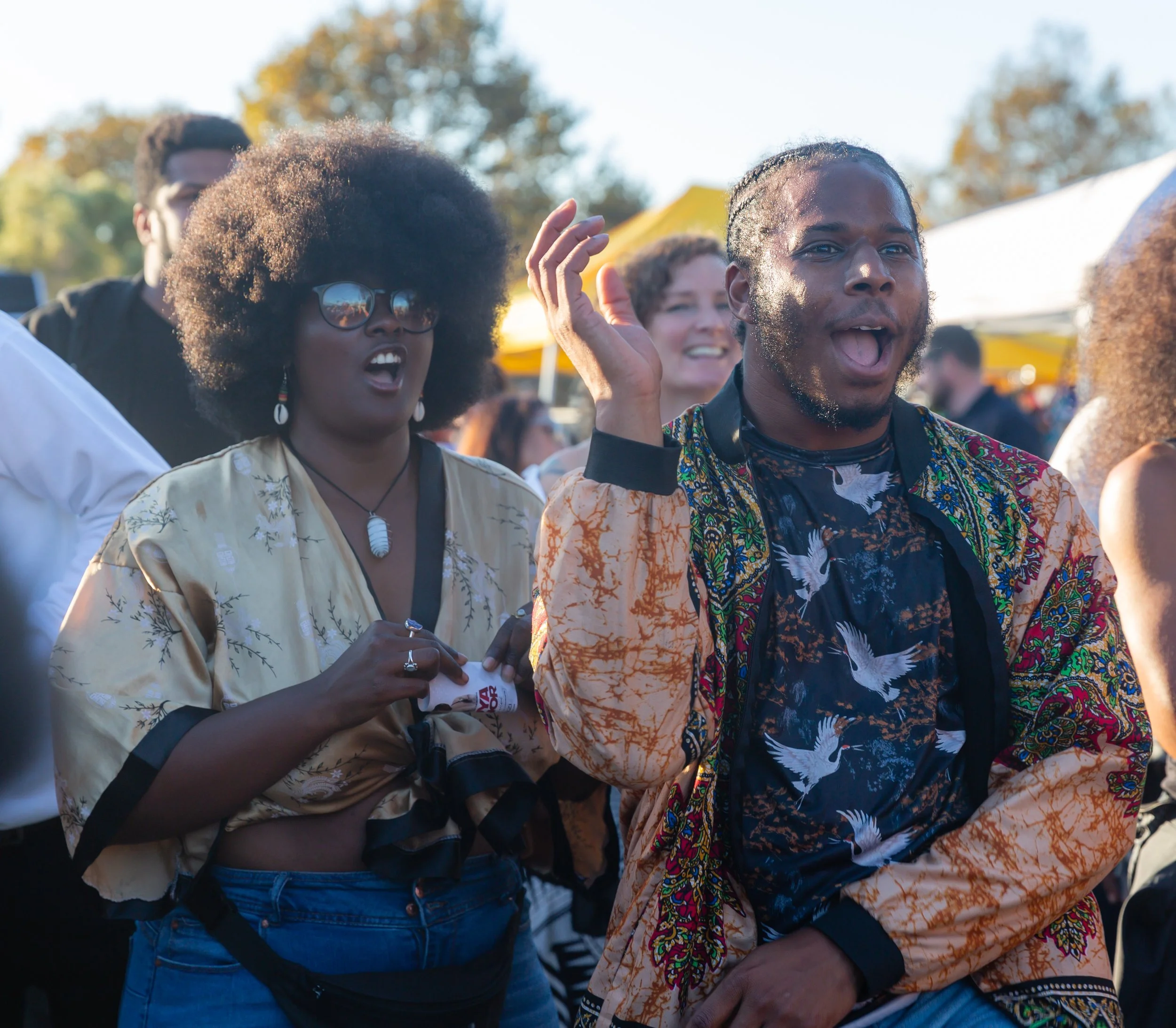 Group of diverse people at an outdoor event, smiling and engaging, with trees and tents in the background.