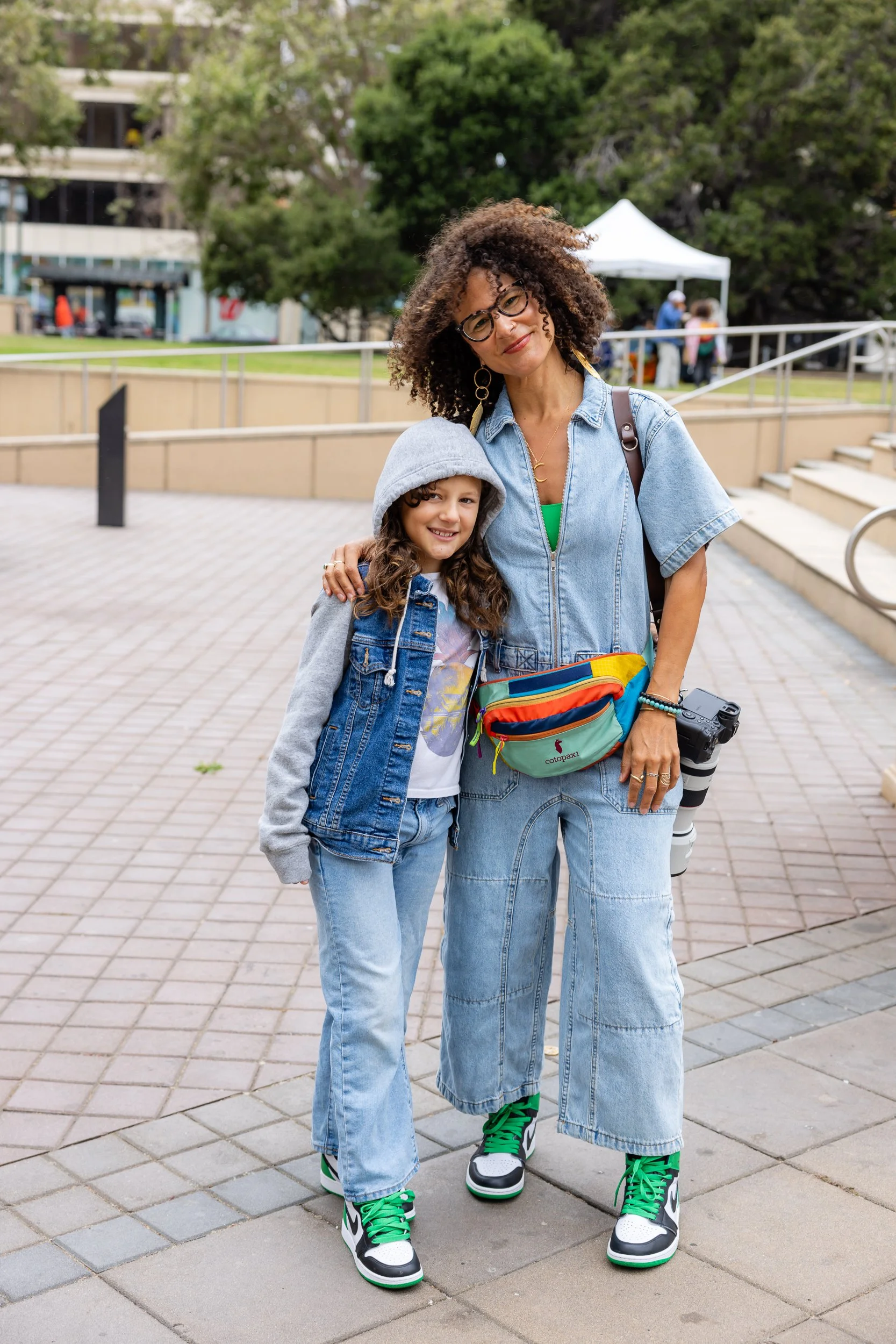 Woman and child standing outdoors on a paved walkway, with trees and a white tent in the background. The woman is wearing glasses, a denim jacket, and colorful sneakers. The child is dressed in a hooded sweatshirt, denim jacket, and has curly hair. They are smiling and posing for the camera.