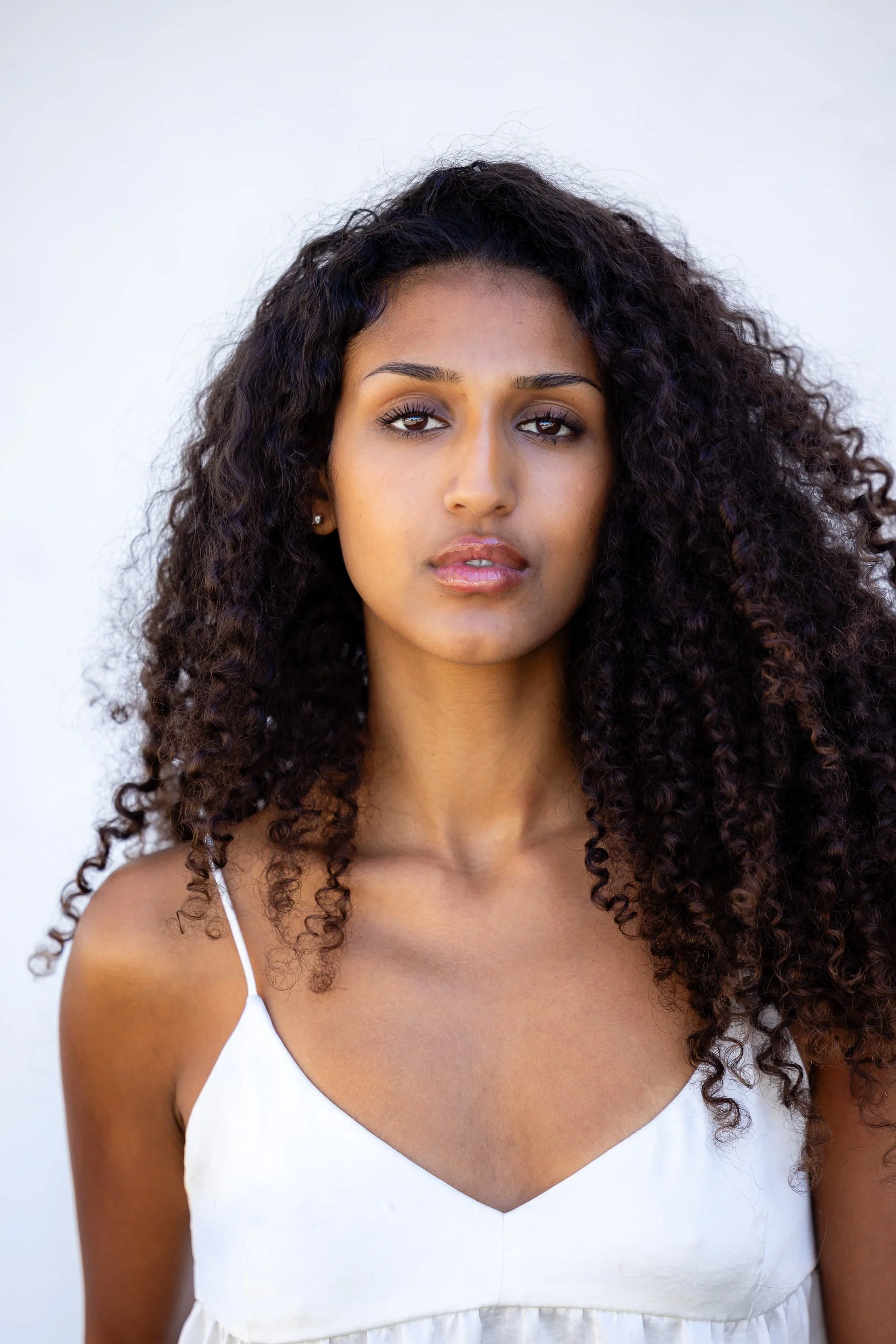 A woman with long, curly dark hair wearing a white sleeveless top against a plain light background.