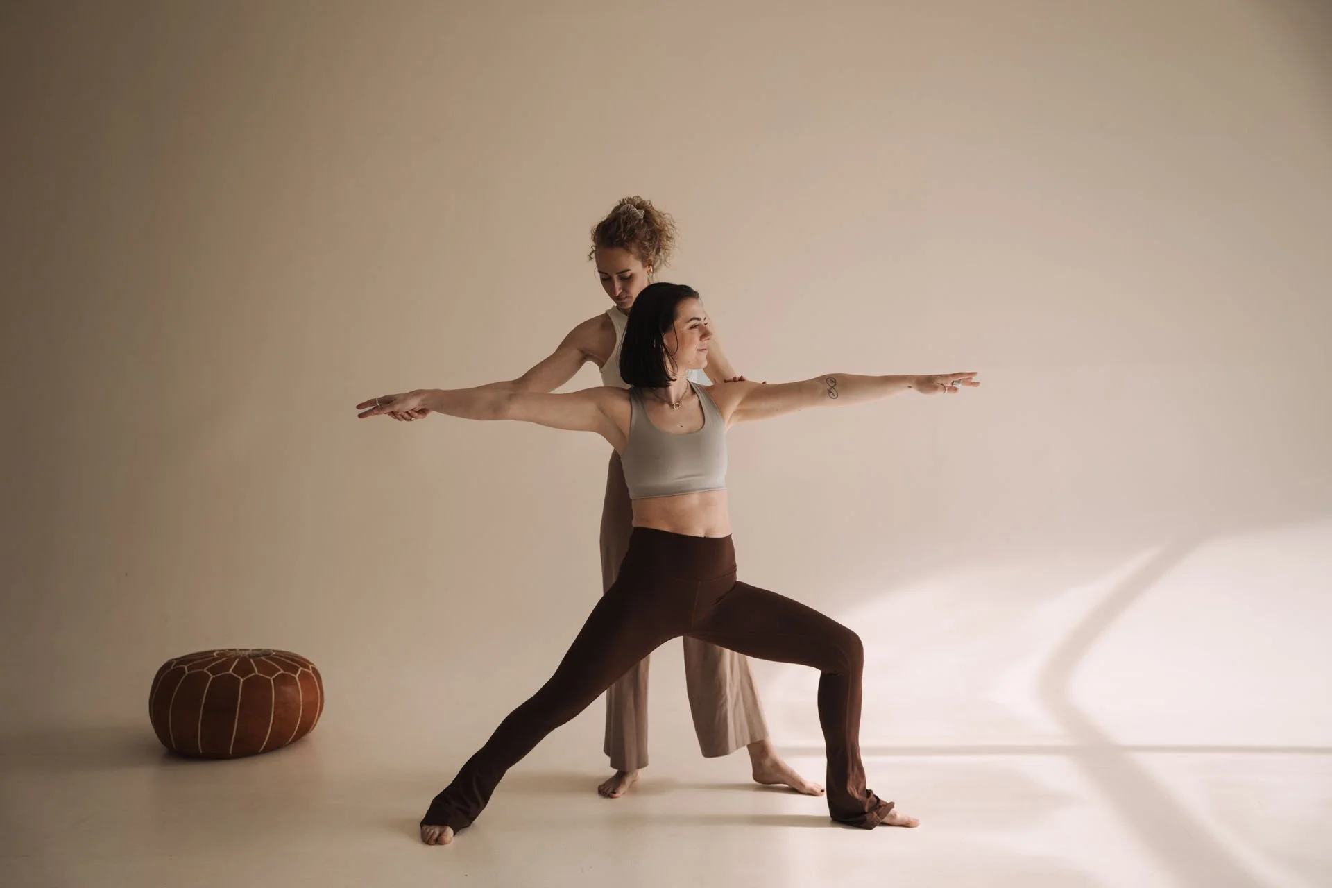 Zwei Frauen praktizieren Yoga in einer neutralen Umgebung, eine in einer Kriegerpose, die andere unterstützt sie im Hintergrund.