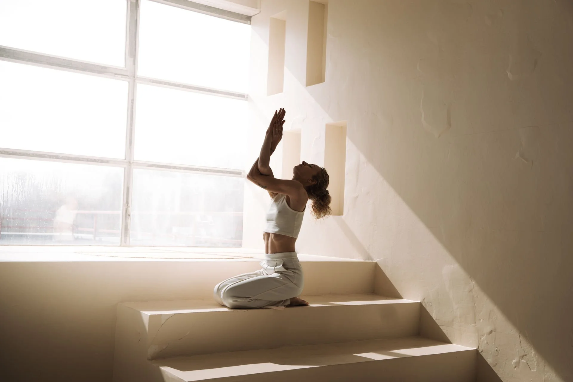Frau in weißen Yoga clothing macht eine Yoga-Übung in einem Raum mit großen Fenstern und natürlichem Licht.