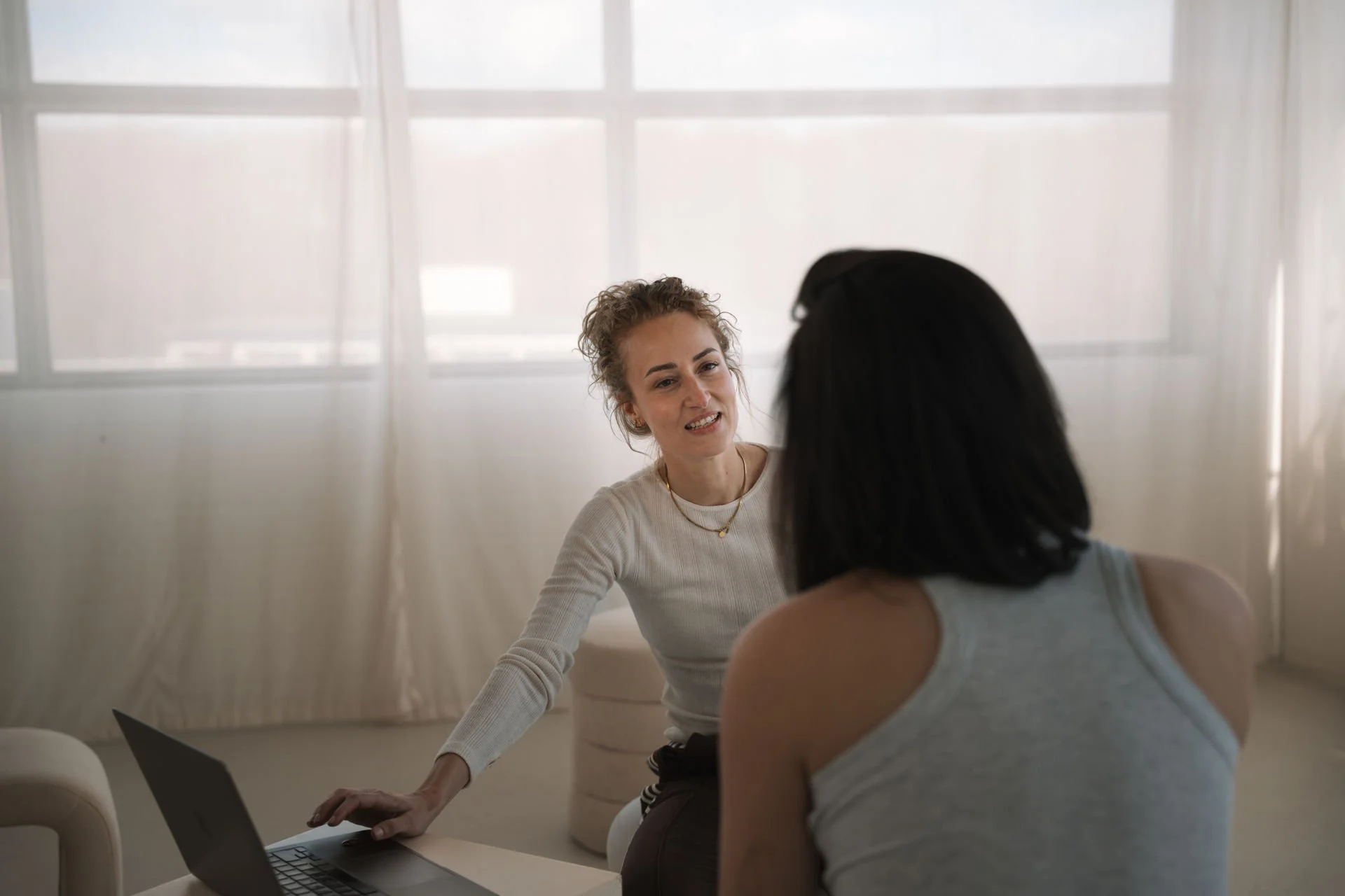 Zwei Frauen unterhalten sich in einem hellen Raum, eine sitzt an einem Tisch mit einem Laptop, die andere steht neben ihr.