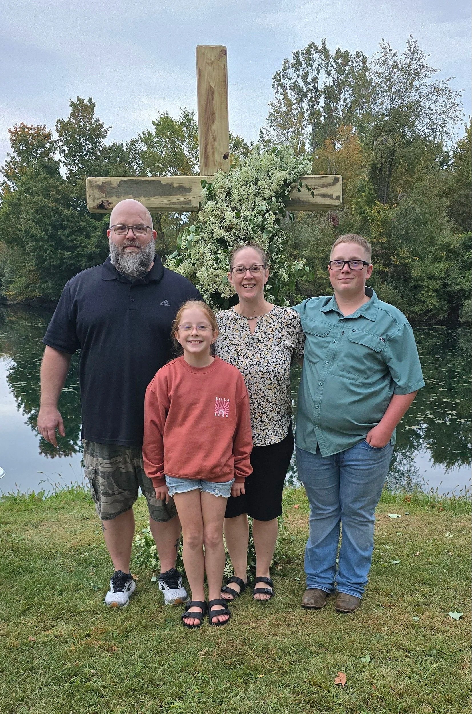 Family of four standing outdoors near a river with a wooden cross decorated with flowers in the background.