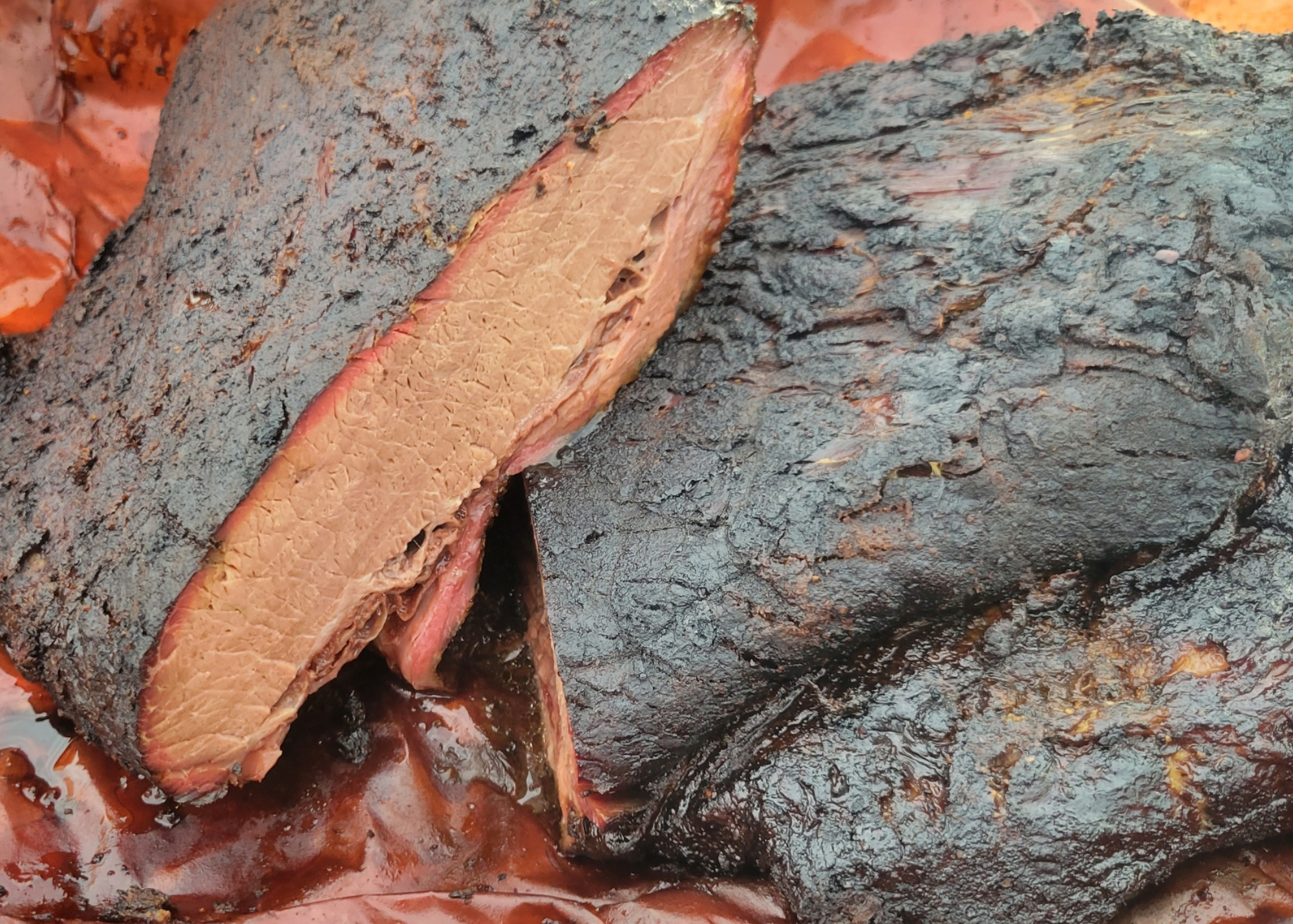 Close-up of a smoked brisket with a dark, bark crust, partially sliced to show the tender, pink smoke ring inside, resting in its juiciness.