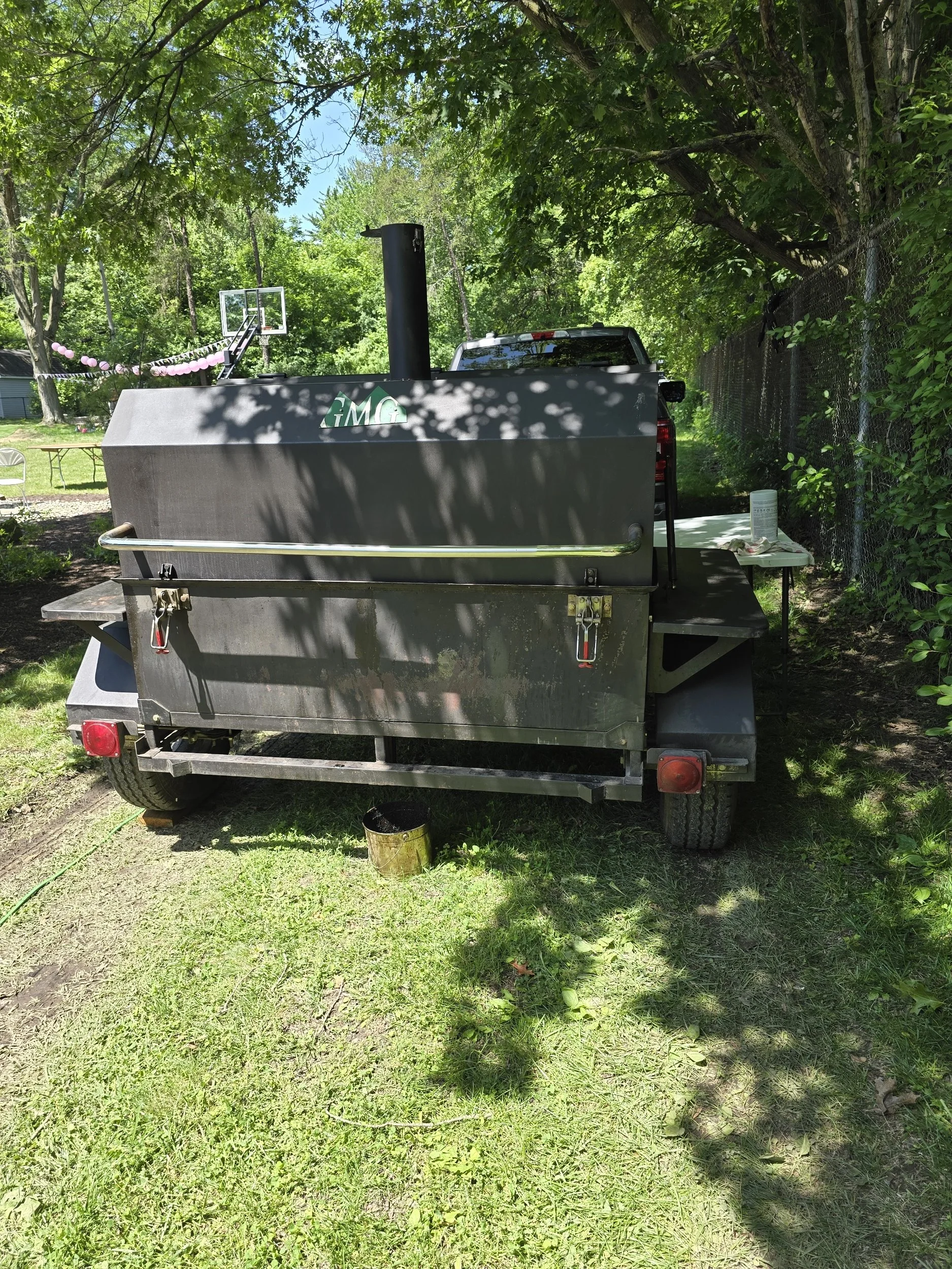 Propane smoker grill on a grassy area with trees, a chain-link fence, a table with a container and paper, a pickup truck, a basketball hoop, and hanging pink balloons in the background.