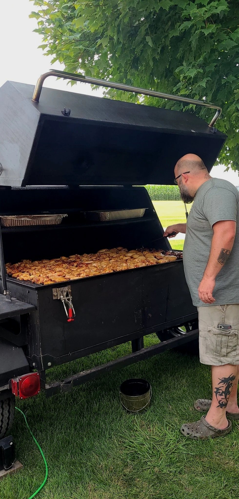 A man with a tattoo on his leg and glasses standing next to a large outdoor smoker grill, cooking a large batch of chicken wings, in a grassy area with trees in the background.