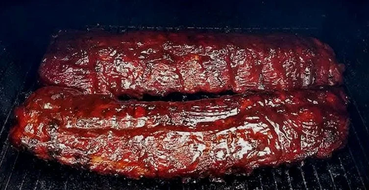 Two racks of BBQ ribs cooking on a grill with a shiny, glazed surface.