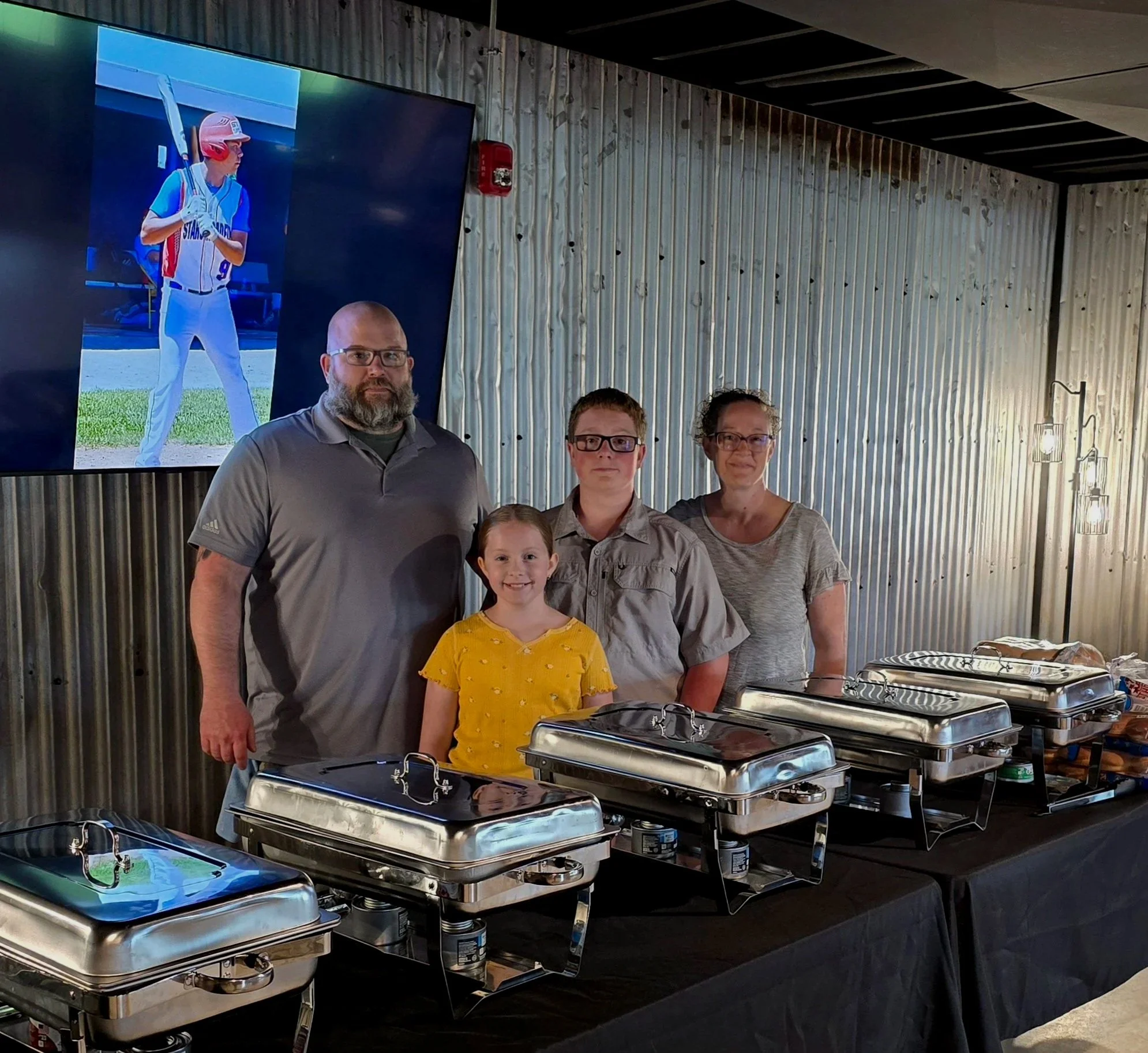 Family standing in front of a buffet table with chafing dishes, inside a venue with corrugated metal walls and a large television screen showing a young baseball player in uniform.