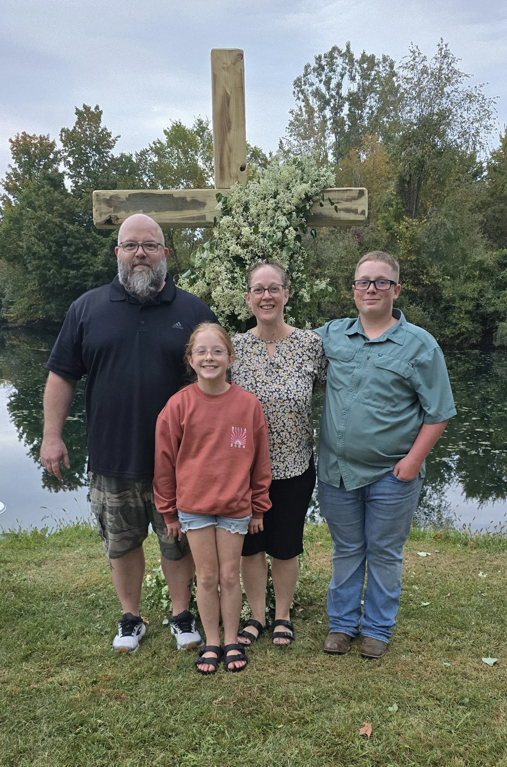 A family of four standing outdoors near a river with a large wooden cross decorated with flowers behind them. The family includes a man, woman, young girl, and teenage boy. They are smiling and posing for the photo, with trees in the background.