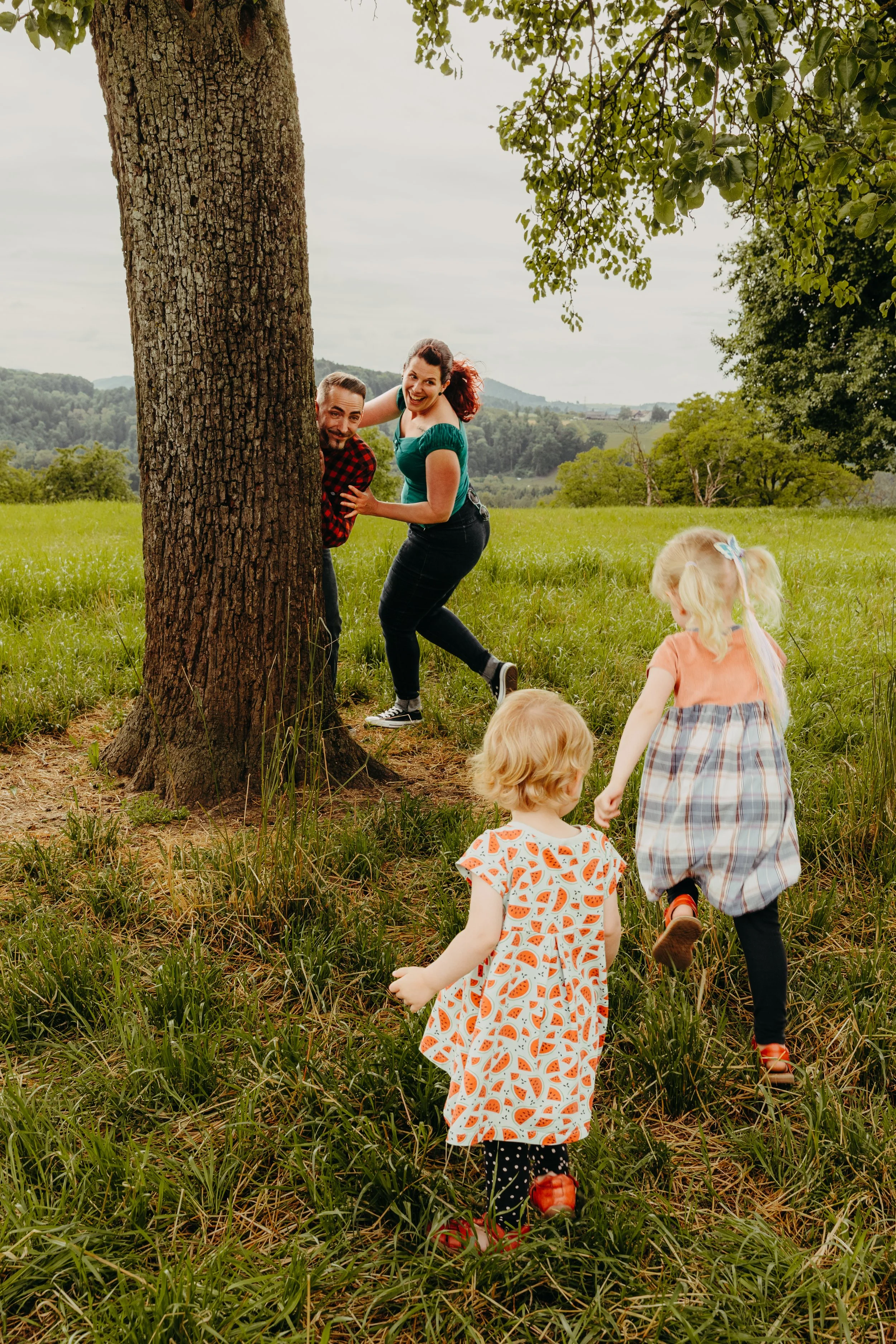 Kinder und Erwachsene spielen im Freien in einem grünen Feld unter einem Baum, das Mädchen trägt ein oranges Kleid mit Wassermelonenmuster, die Erwachsenen verstecken sich hinter einem Baum, während die Kinder barfuß laufen.