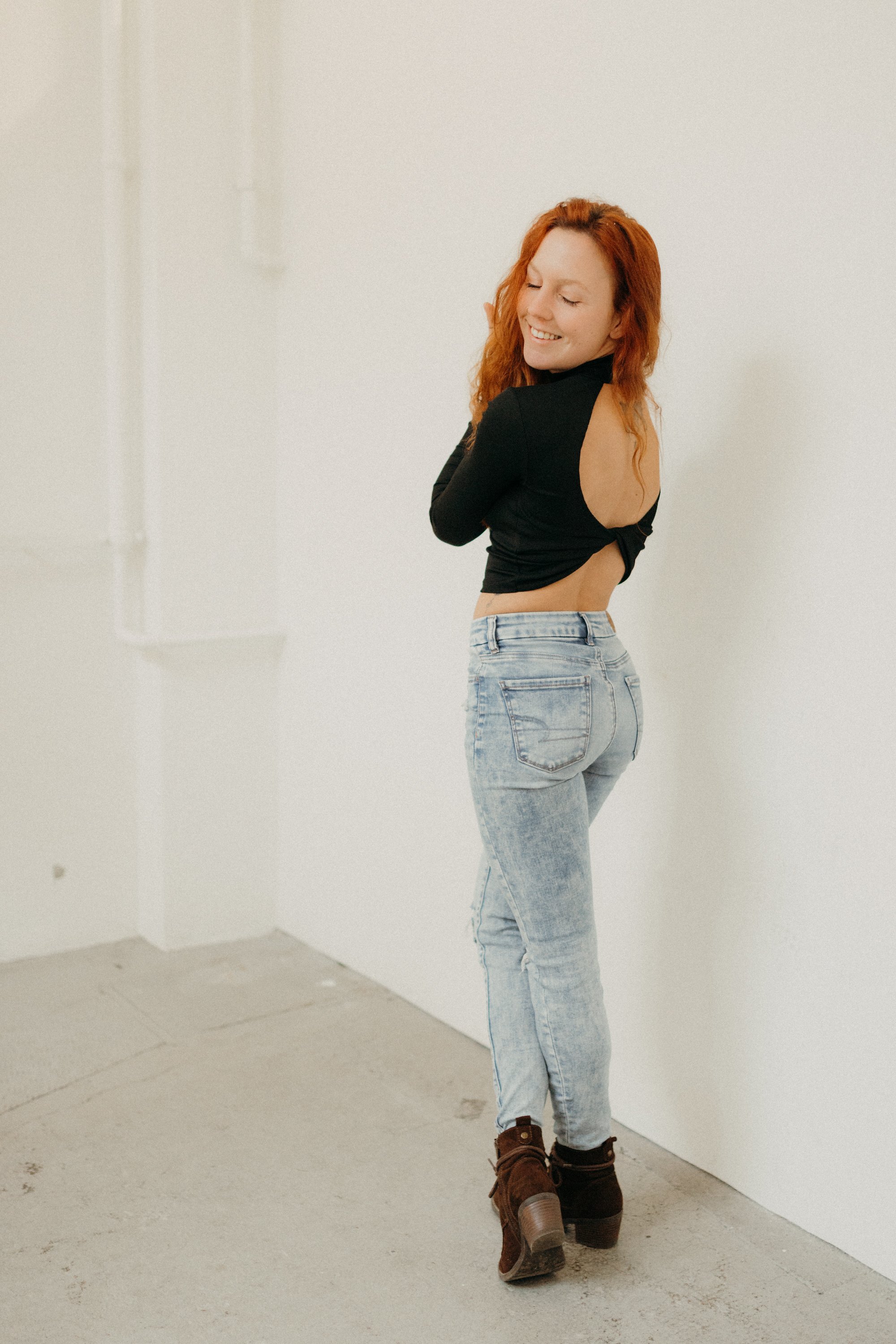 Eine junge Frau mit roten, lockigen Haaren trägt eine schwarze Jacke mit einem offenen Rücken, hellblaue Jeans und braune Stiefel. Sie steht in einem weißen Raum, lächelt und dreht sich leicht zur Seite.