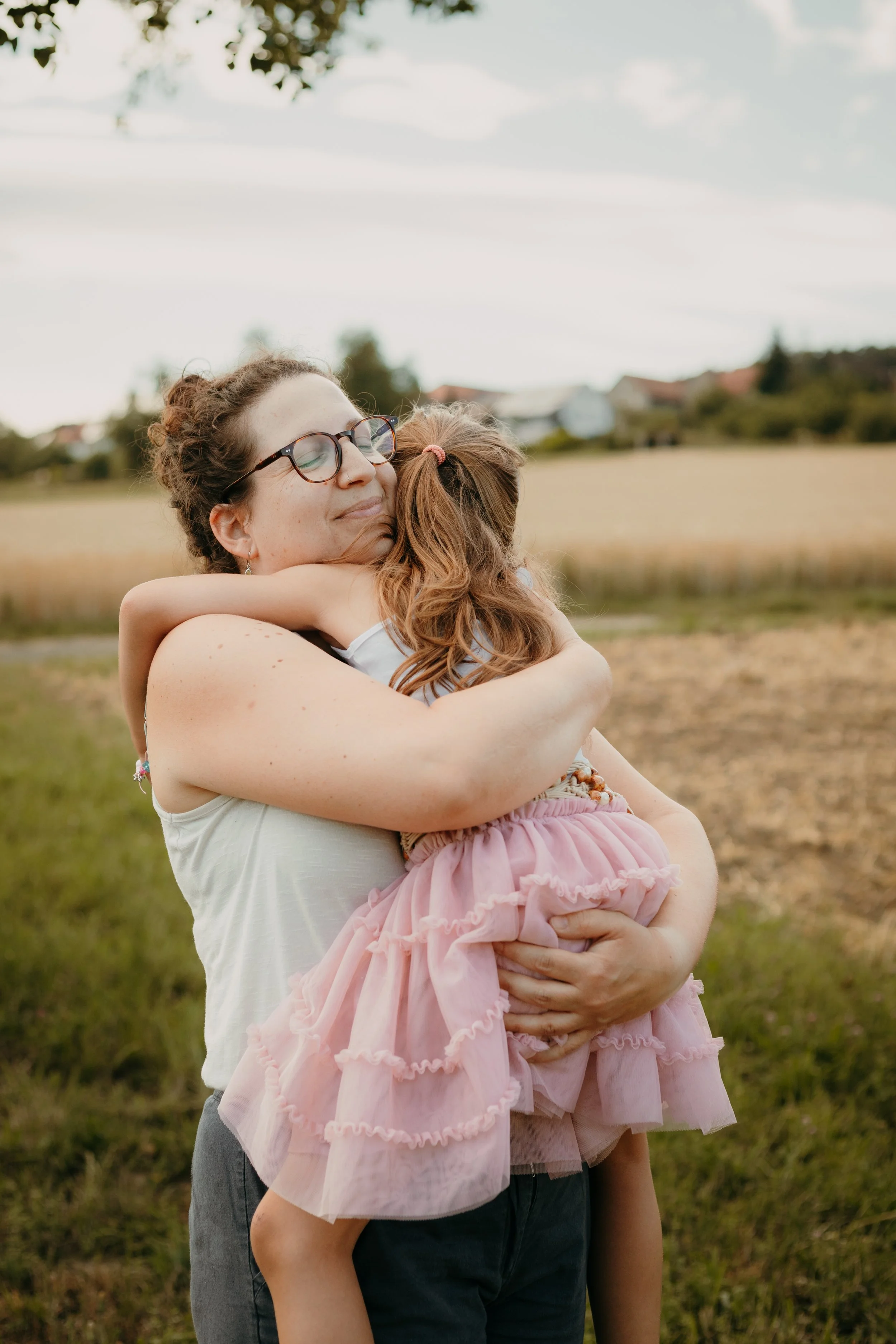 Eine Frau umarmt ein Kind in einem Feld, beide lächeln. Es ist ein naturnaher Hintergrund mit Häusern im Hintergrund.
