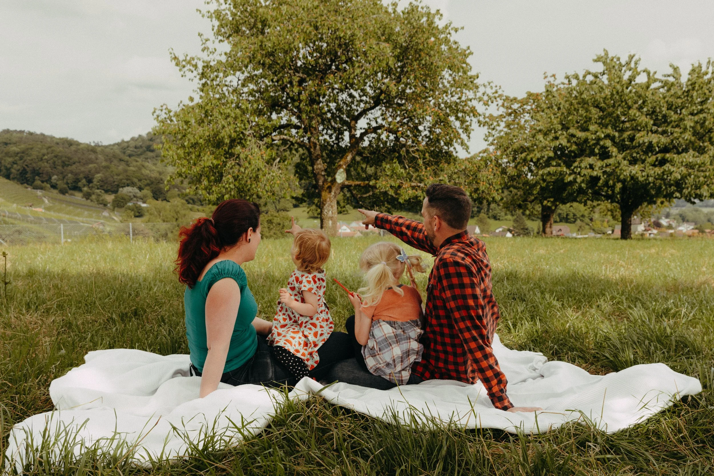 Familie sitzt auf einer weißen Decke im Grünen und zeigt auf einen Baum im Hintergrund.