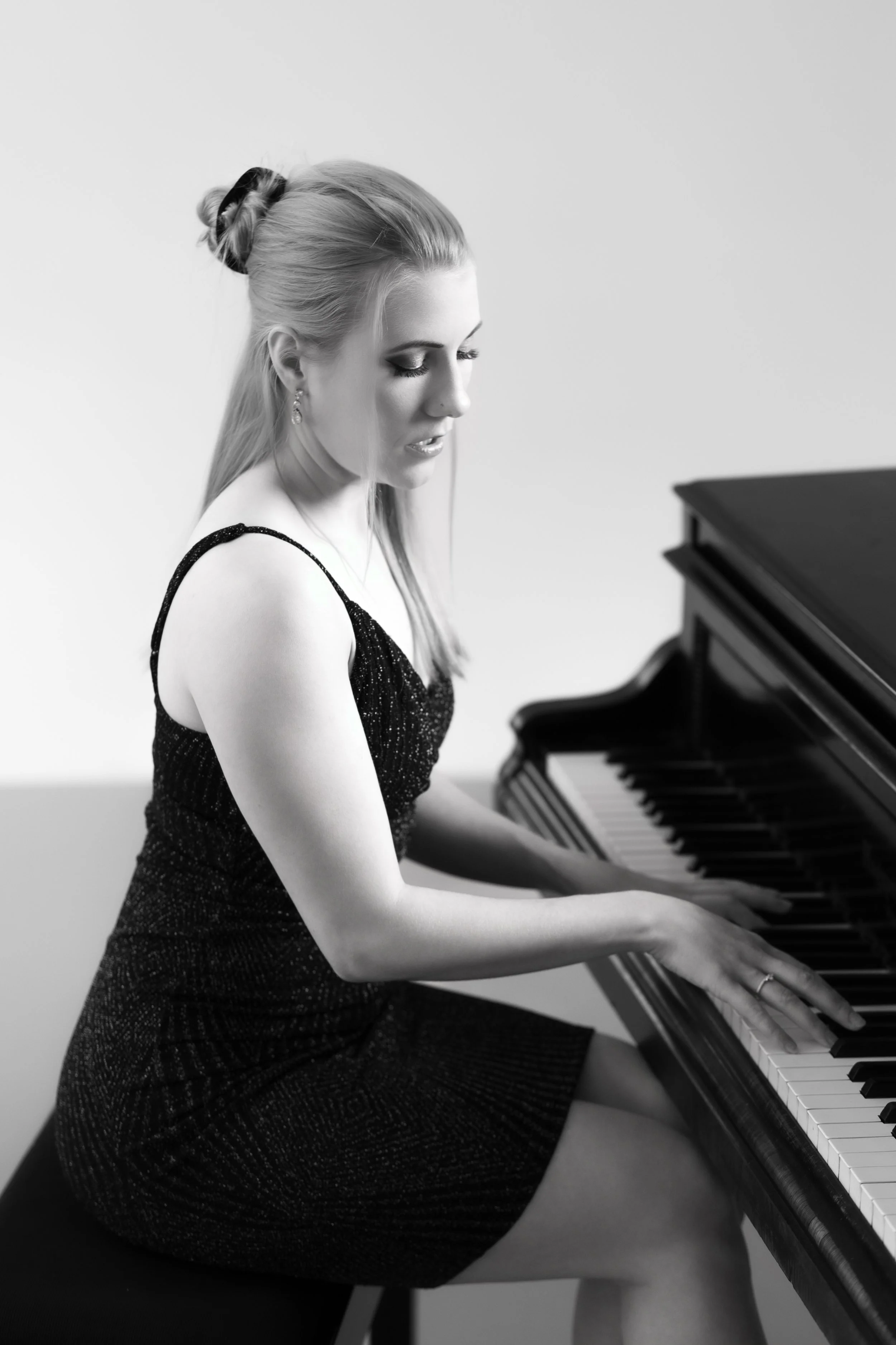Schwarzweißfoto einer jungen Frau, die an einem Klavier spielt, mit hochgestecktem Haar und eleganter Kleidung, in einem schlichten Raum.