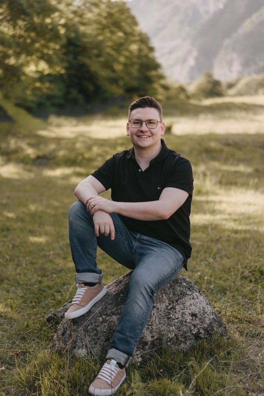 Ein glücklicher junger Mann sitzt auf einem großen Stein in einer natürlichen, grünen Landschaft mit Wasser und Bäumen im Hintergrund.