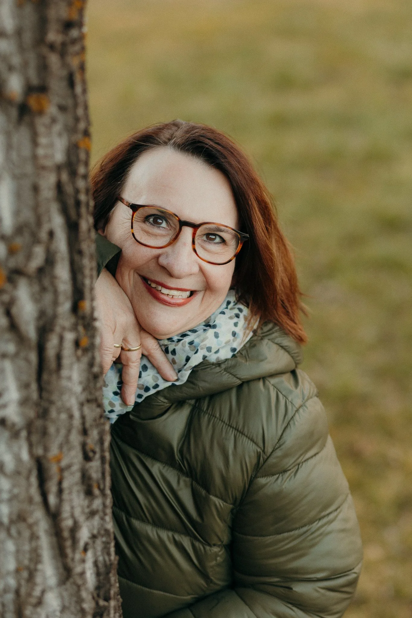 Eine lachende Frau mit Brille, die hinter einem Baum versteckt ist, im Freien im Grünen.