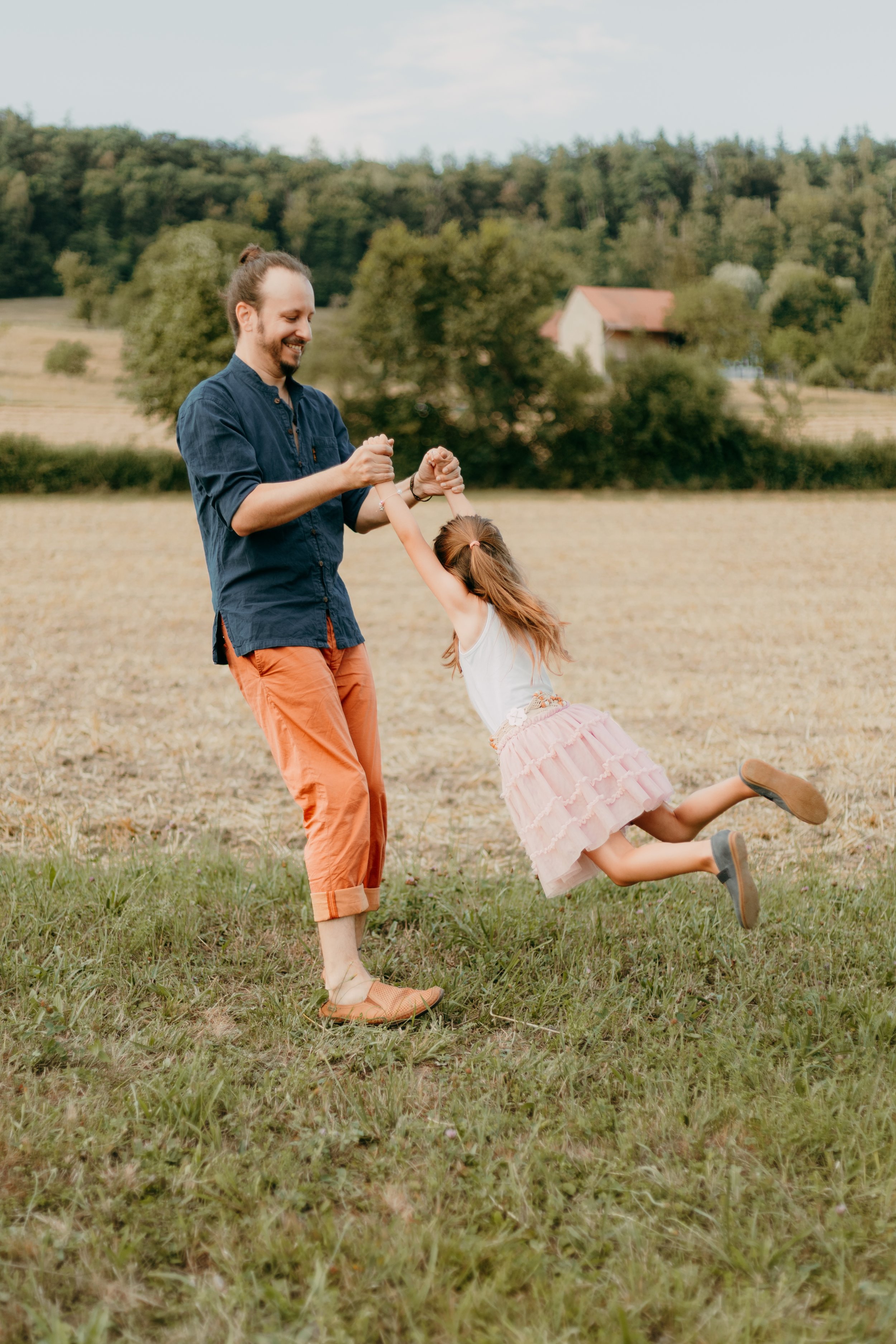 Ein Mann hält ein Mädchen in einem grasbewachsenen Feld, sie spielen. Im Hintergrund sind Bäume und ein Haus mit rotem Dach.