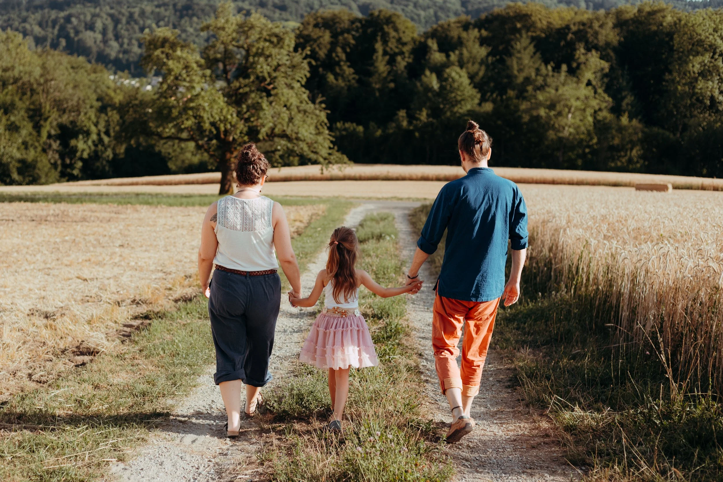 Eine Familie mit zwei Erwachsenen und einem Kind, die einen Feldweg entlang in die Natur gehen, umgeben von Feldern und Bäumen bei Sonnenuntergang.