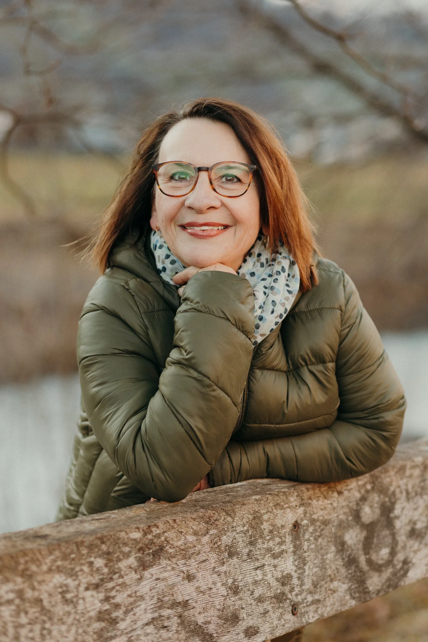 Eine lachende Frau mit Brille und roten Haaren trägt einen kurzen Parka und eine gepunktete Halskette. Sie sitzt an einem Baumstamm im Freien, im Hintergrund sind unscharfe Bäume und Natur zu sehen.