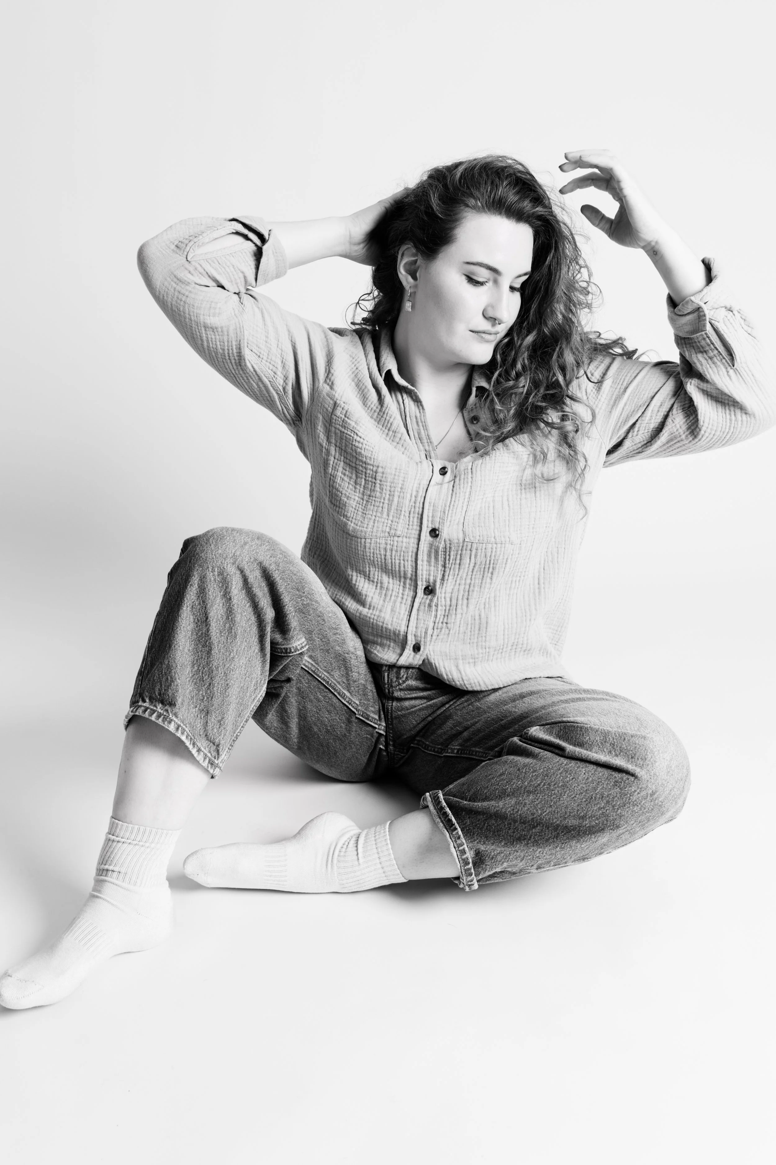 Schwarz-Weiß-Foto einer jungen Frau mit lockigem Haar, die im Sitzen am Boden entspannt ist, mit Blick nach unten, trägt ein lässiges Hemd, Jeans und Turnschuhe.