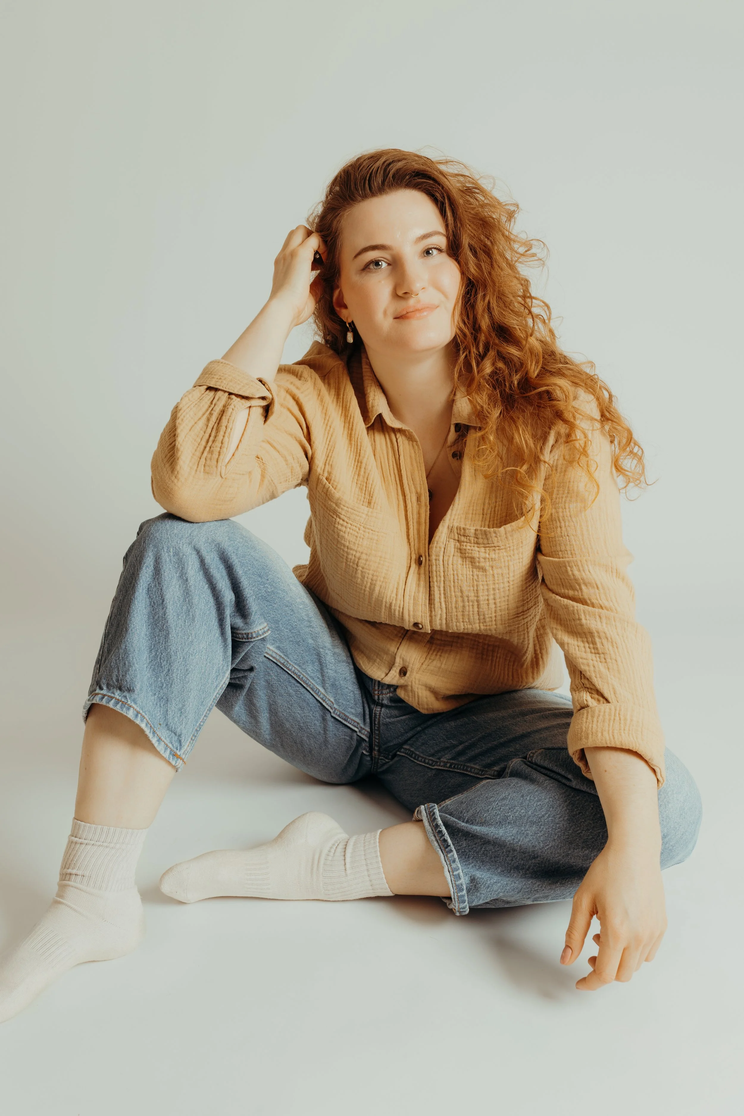 Eine junge Frau mit lockigem roten Haar sitzt auf dem Boden, trägt eine beige Cordjacke, blaue Jeans und weiße Socken, posiert lächelnd vor neutralem Hintergrund.