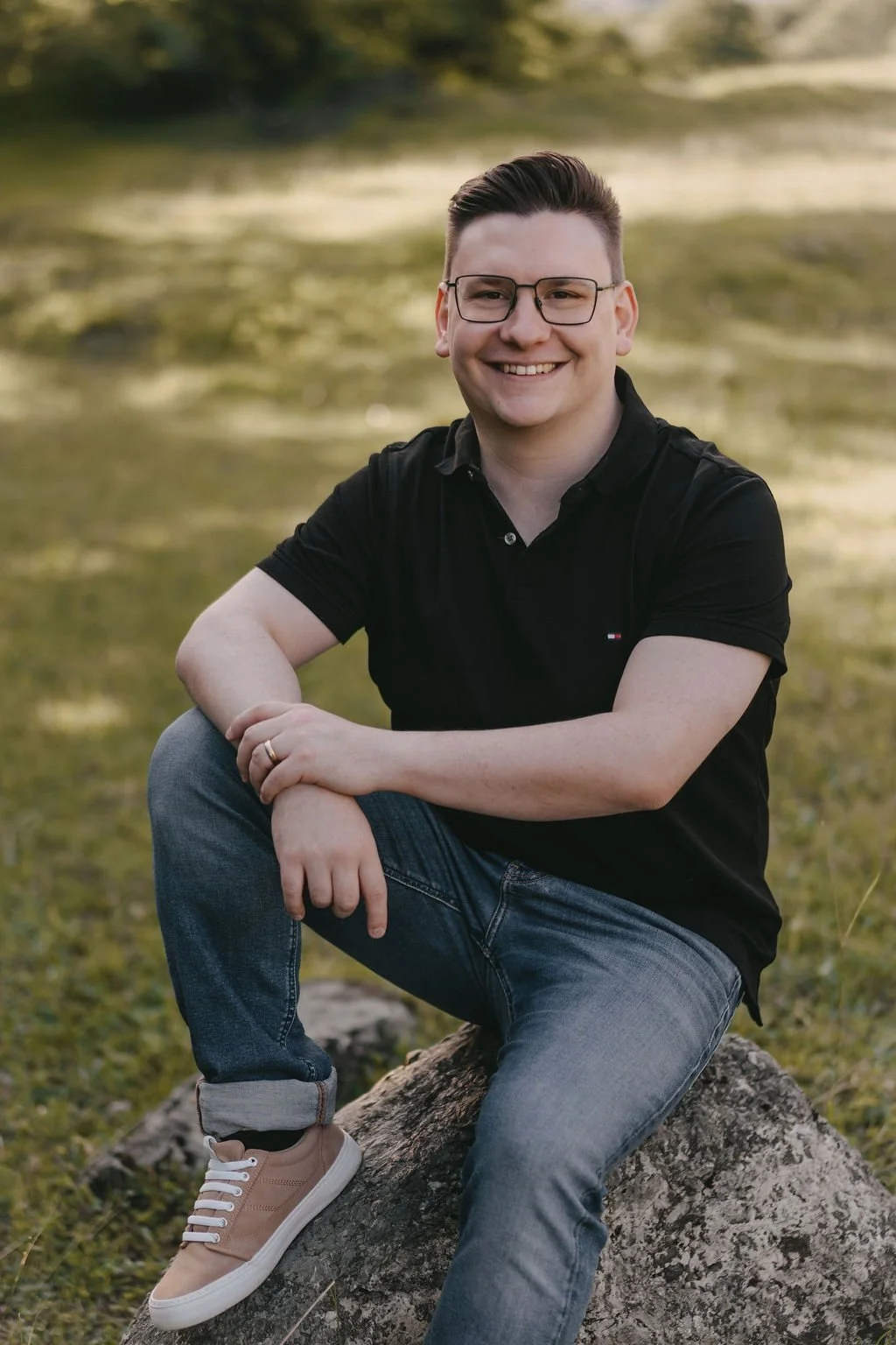 Ein Mann sitzt auf einem Baumstamm im Freien, trägt eine schwarze Polo-Hemd, Jeans und Sneakers, lächelt in die Kamera.