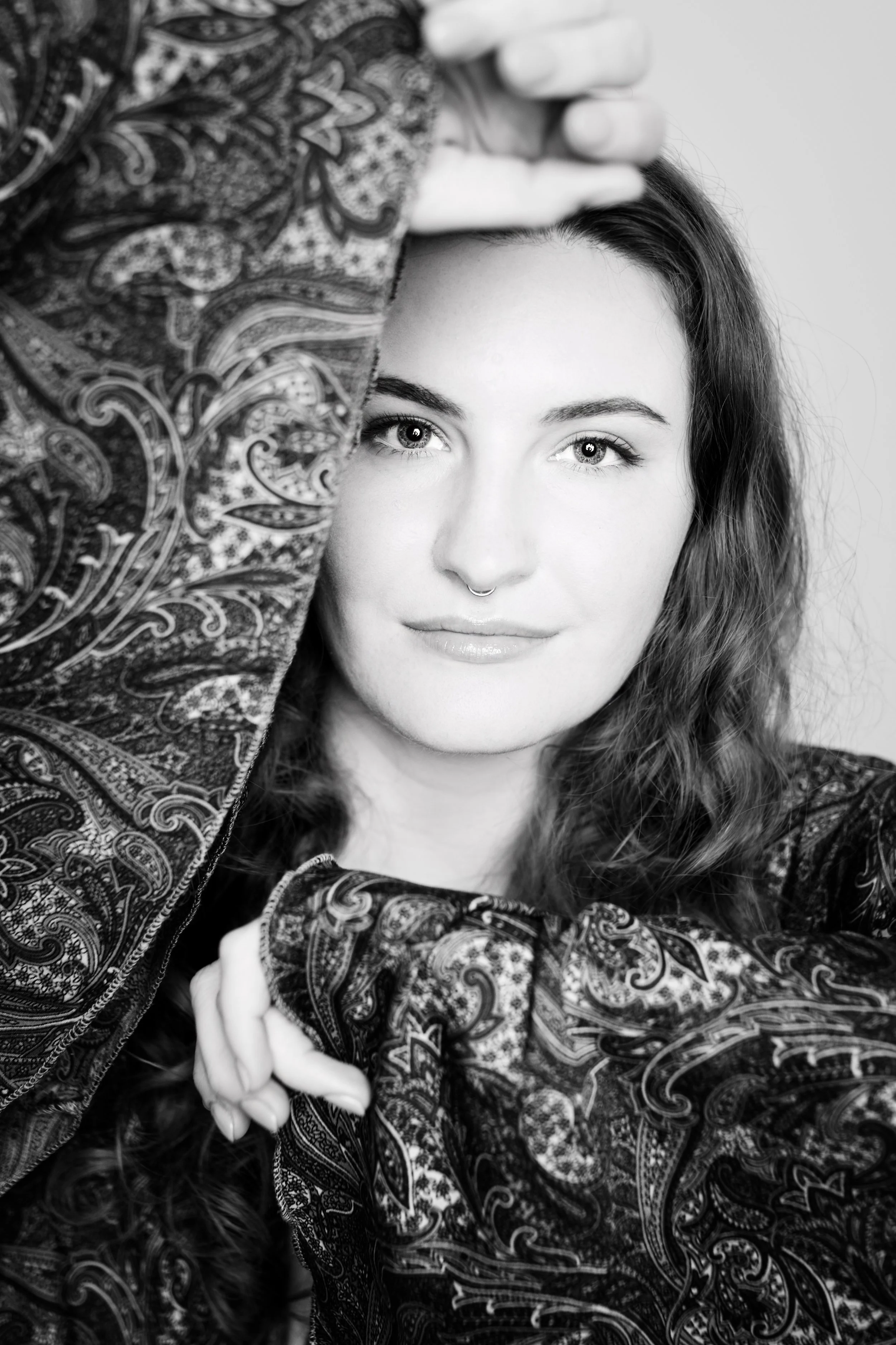 Schwarz-weiß-Fotografie eines jungen Frauenporträts, bei dem ihre Augen im Fokus stehen. Sie hat mittellanges, lockiges Haar, eine Nasenpiercing und trägt ein Oberteil mit einem detaillierten Paisleymuster. Ihre Hand ist sichtbar, während sie eine Rolle spielt oder eine Pose einnimmt.