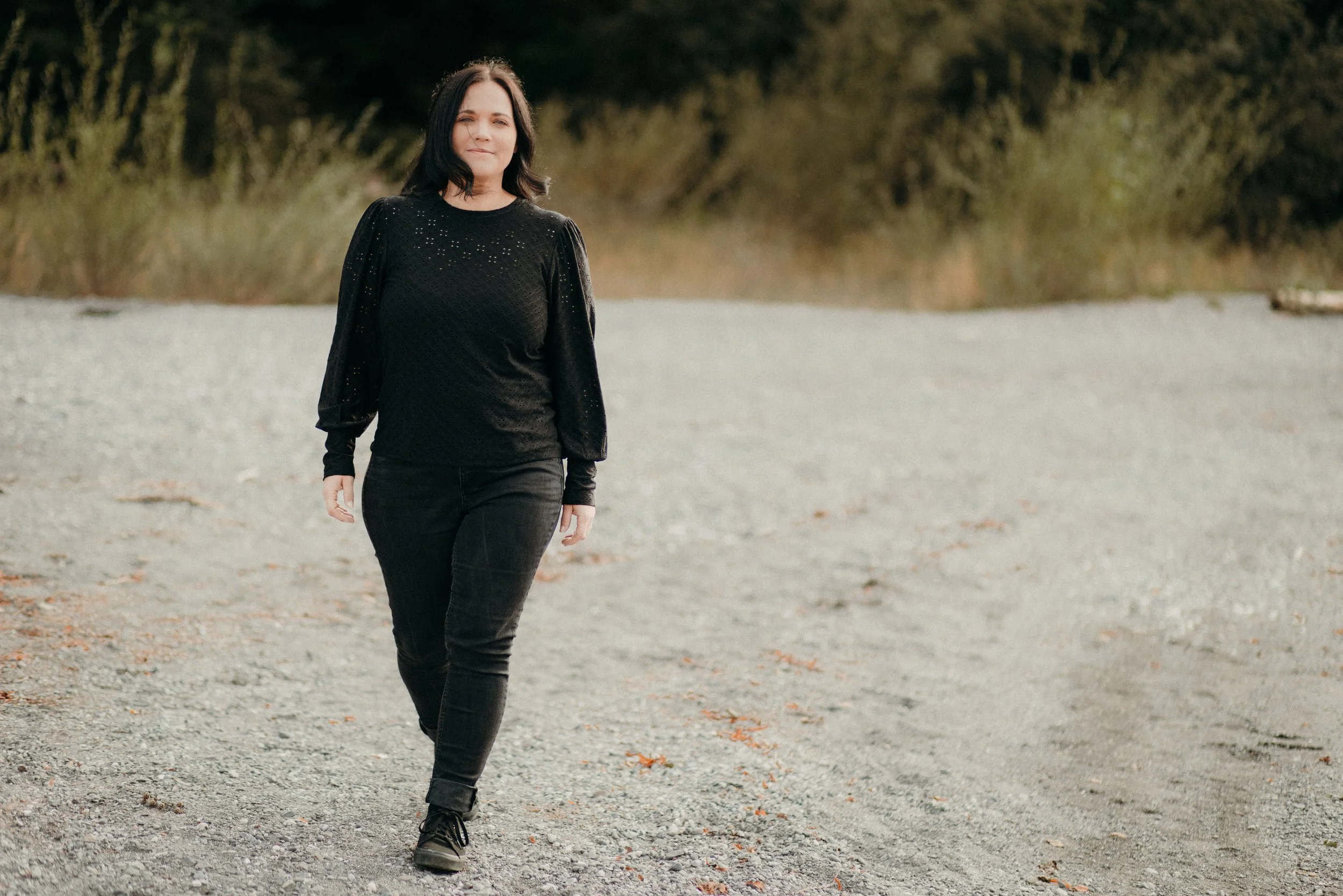 Eine Frau mit schwarzer Haarfarbe, schwarzem Pullover mit Lochmustern und schwarzen Hosen, spaziert auf einem Kiesweg im Freien, im Hintergrund Bäume und Büsche, bei Tageslicht.