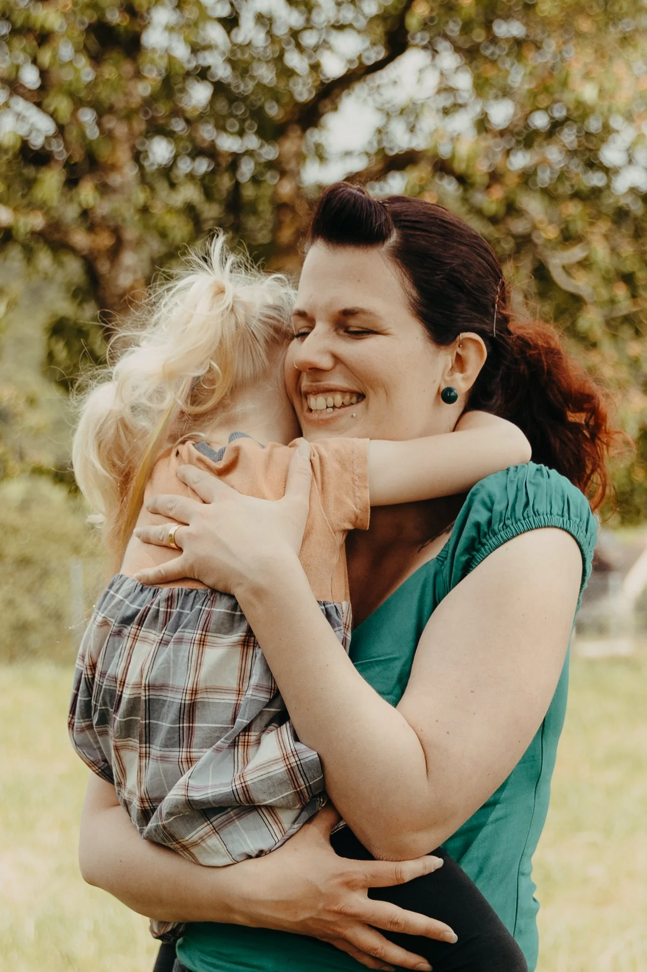 Eine Frau mit dunklen Haaren und Piercings umarmt und hält ein kleines blondes Mädchen im Park. Die beiden lachen und zeigen Zuneigung, im Hintergrund Bäume mit grünen Blättern.