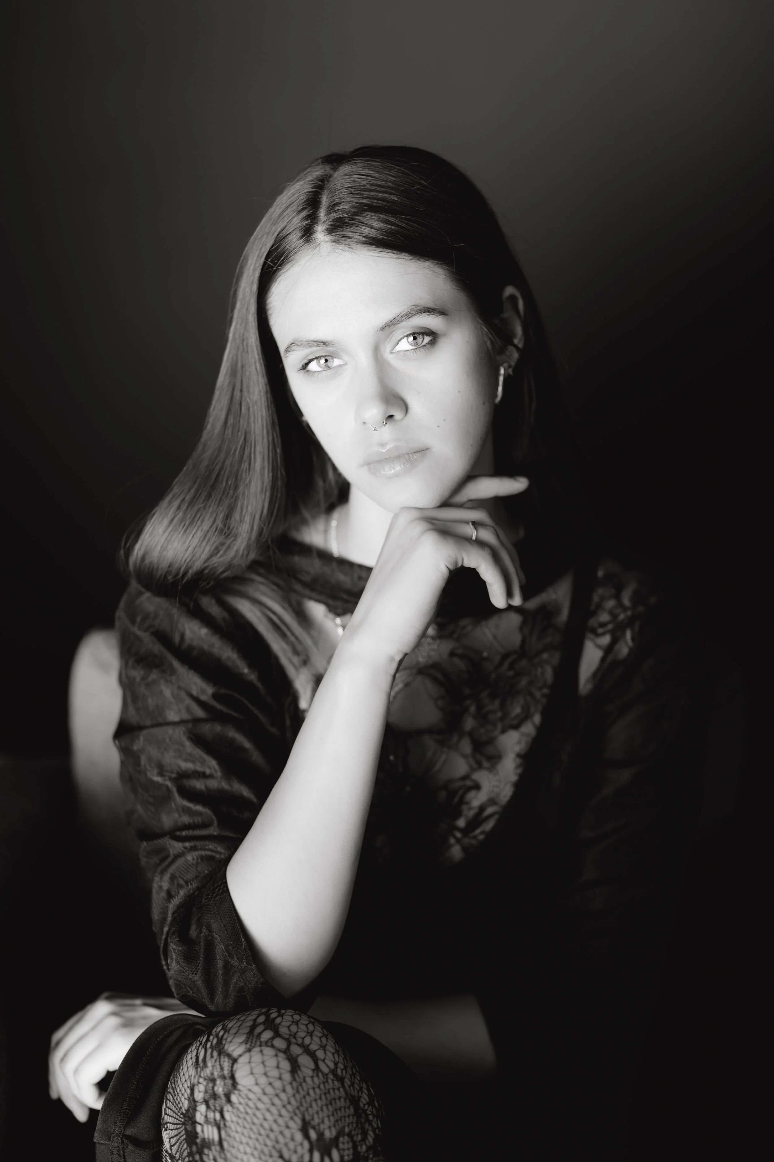 Schwarz-weiß-Fotografie eines jungen Frauenporträts mit langem glatten Haar, trendigem Make-up und Tätowierungen, die nachdenklich wirkt mit Hand am Kinn, sitzend gegen dunklen Hintergrund, elegant gekleidet in Spitzenkleid und Netzstrümpfen.