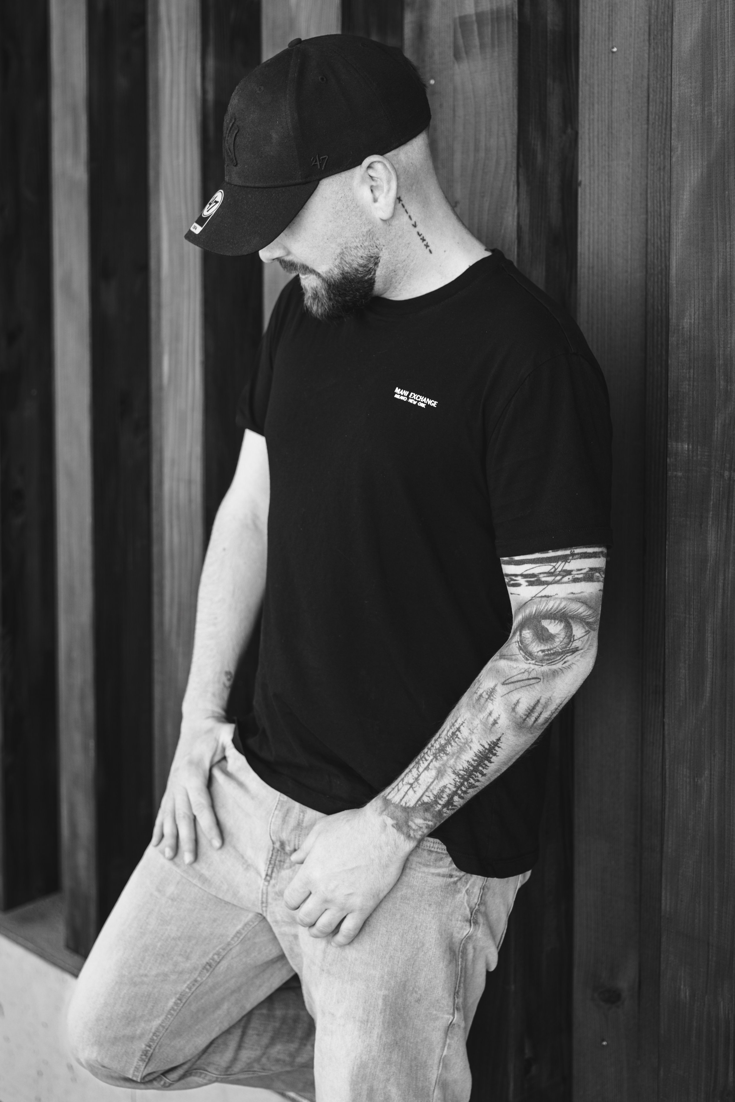 Ein Mann trägt einen schwarzen Baseball-Cap und ein schwarzes T-Shirt, mit Tattoos am Arm, lehnt an einer Holztafel und schaut nach unten.