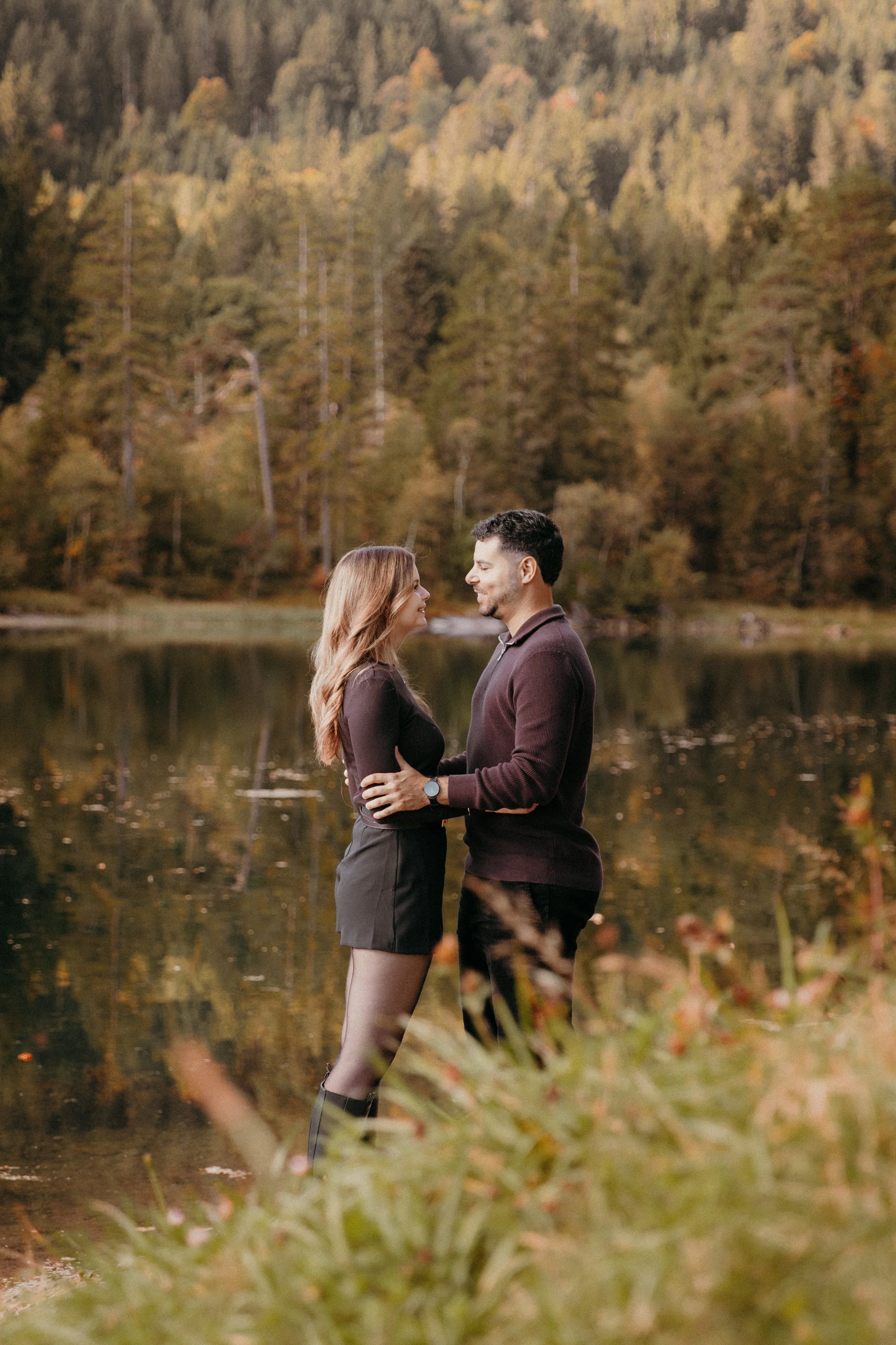 Ein verliebtes Paar steht sich nahe an einem See im Herbst, die Blicke sind in die Augen gerichtet, hinter ihnen bunte Herbstbäume.