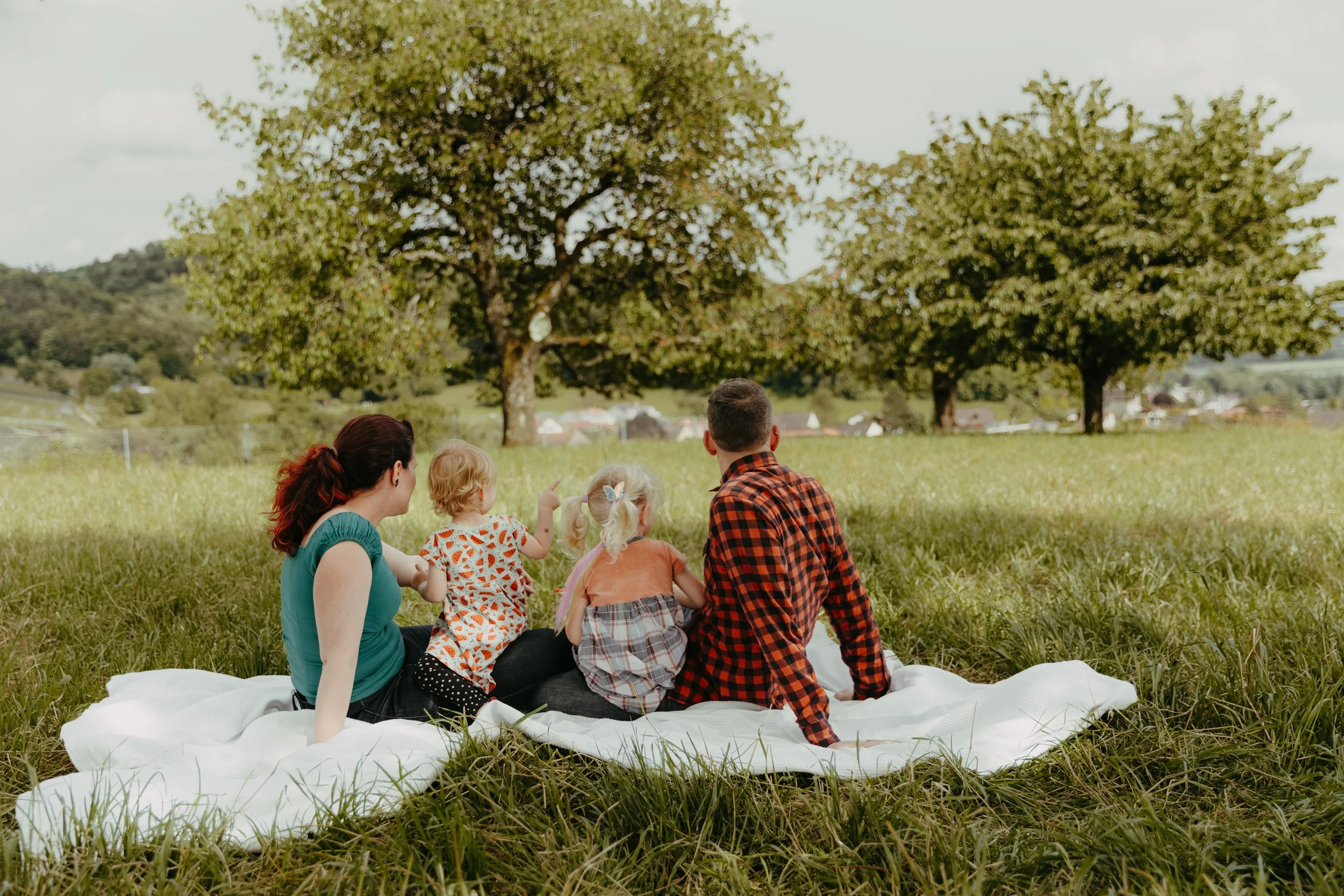 Eine Gruppe von Erwachsenen und Kindern sitzen auf einer weißen Decke im Grünen unter einem Baum, während sie einen Tag im Freien genießen.