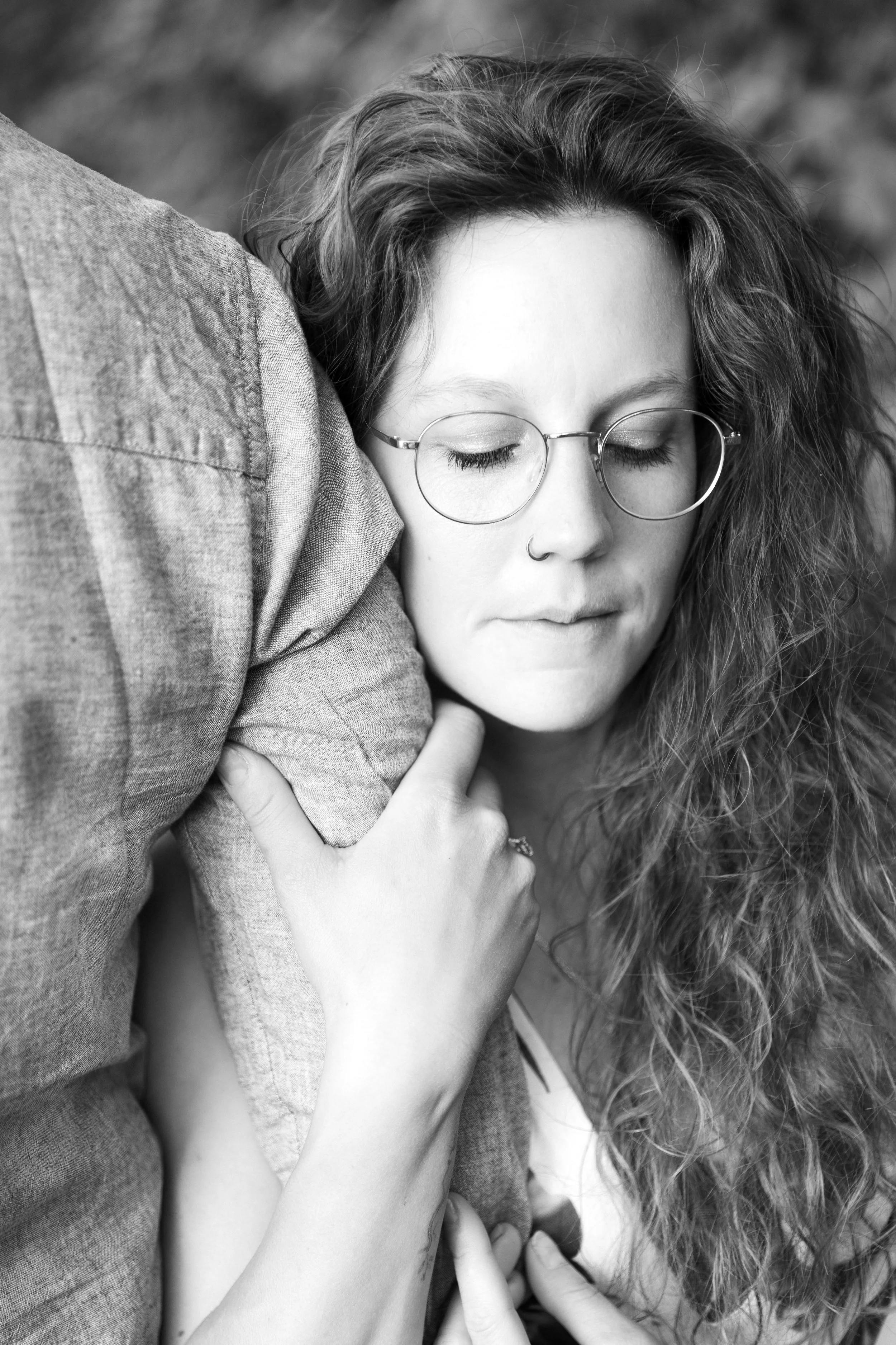 Schwarz-weiß-Foto eines jungen Mädchens mit langen, lockigen Haaren, Brille und Nasenring, das die Augen geschlossen hat, während eine Person die Schulter berührt.