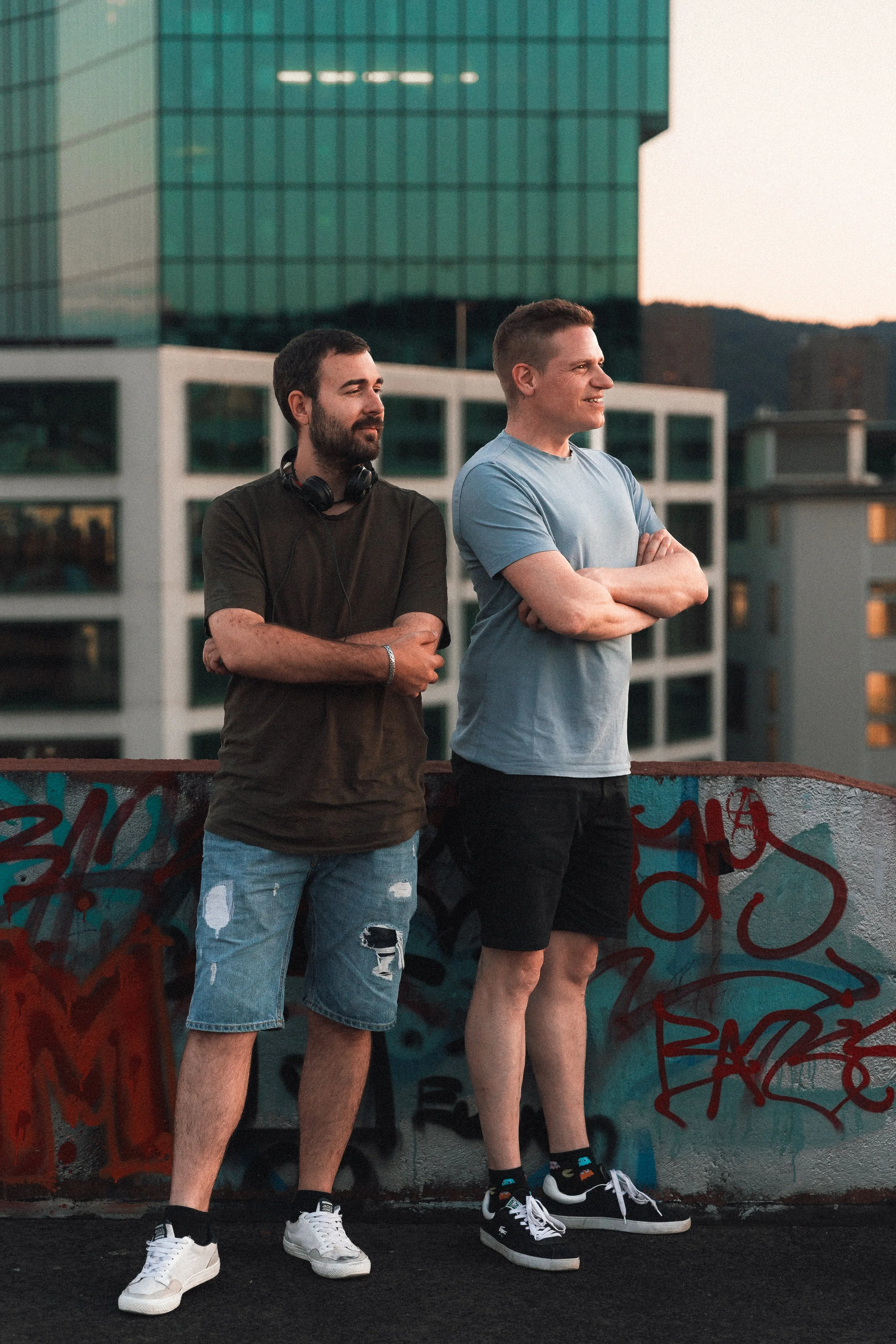 Zwei junge Männer stehen vor einer mit Graffiti bedeckten Wand in einer urbanen Umgebung während des Sonnenuntergangs. Einer trägt ein schwarzes T-Shirt und zerrissene Jeans, der andere ein hellblaues T-Shirt und schwarze Shorts. Beide tragen Turnschuhe.