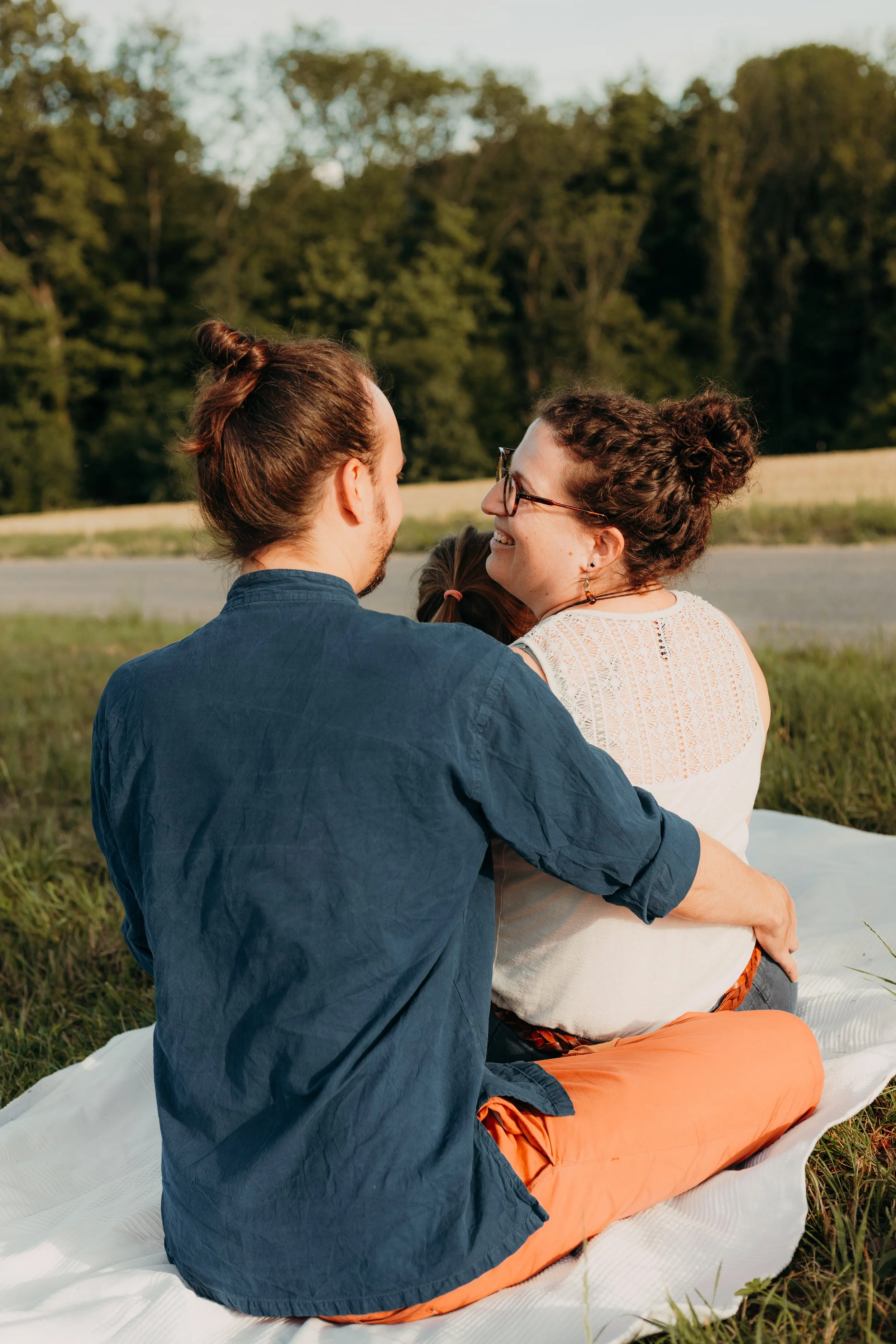 Ein Mann und eine Frau sitzen auf einer weißen Decke auf dem Gras im Freien, die Frau trägt eine weiße Bluse und eine orange Hose, beide lächeln und schauen sich an, im Hintergrund Bäume und Himmel.