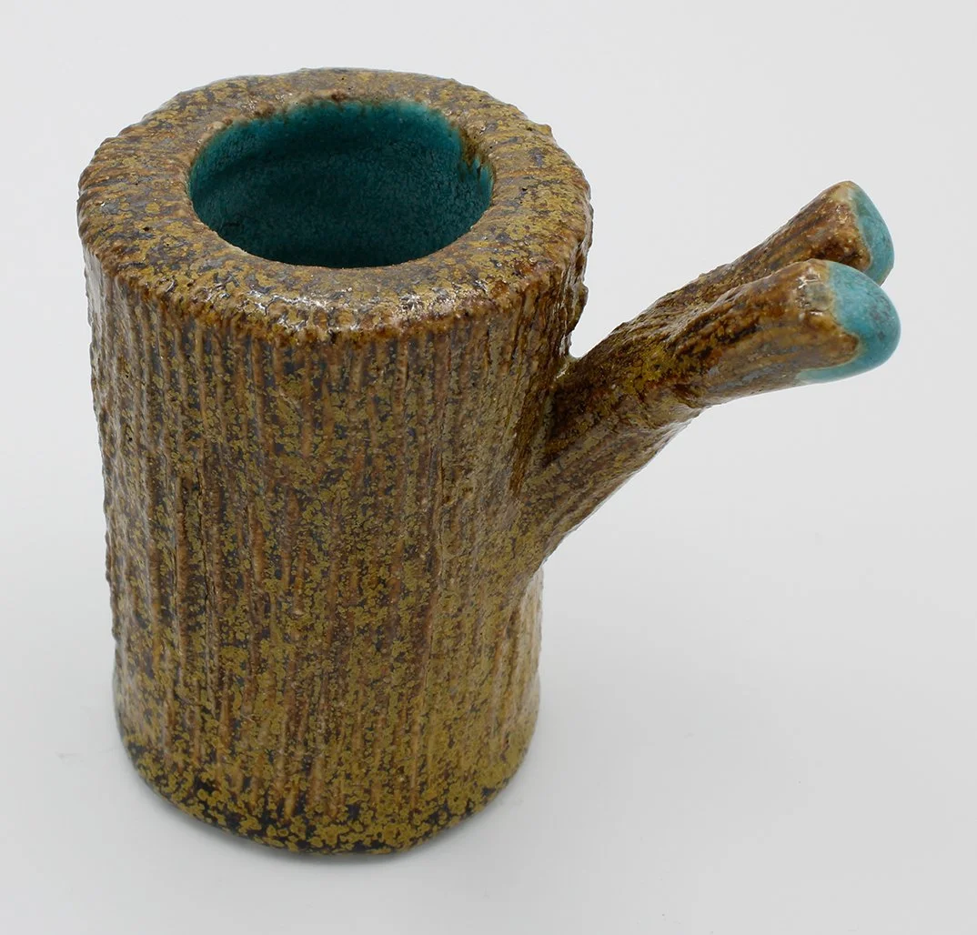 A ceramic mug shaped like a tree trunk with a small branch and turquoise accents at the top of the branch.