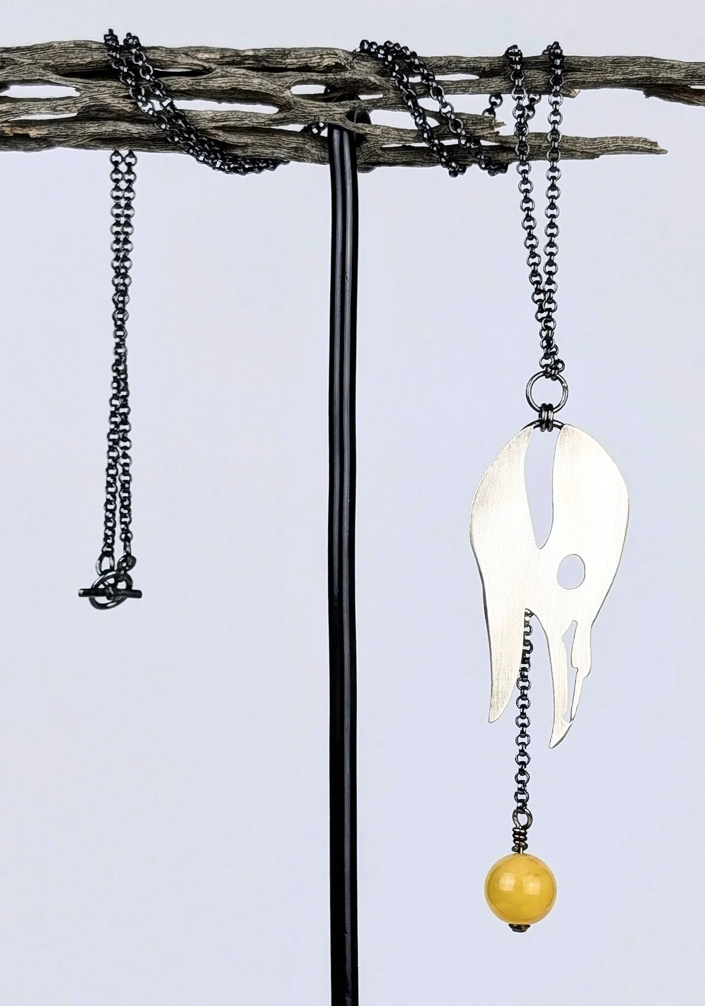 Black necklace chains hanging from a wooden stick with a metallic skull pendant and a yellow spherical pendant.