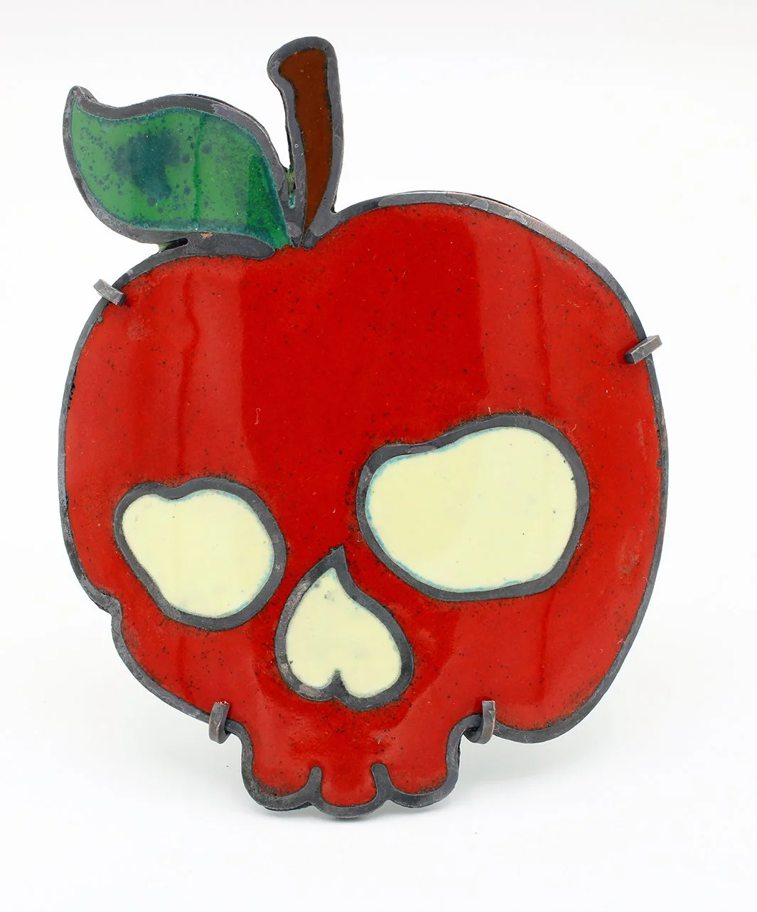 A stained glass art piece in the shape of a red apple with a green leaf and brown stem, featuring skull-shaped cutouts for the eyes, nose, and mouth.