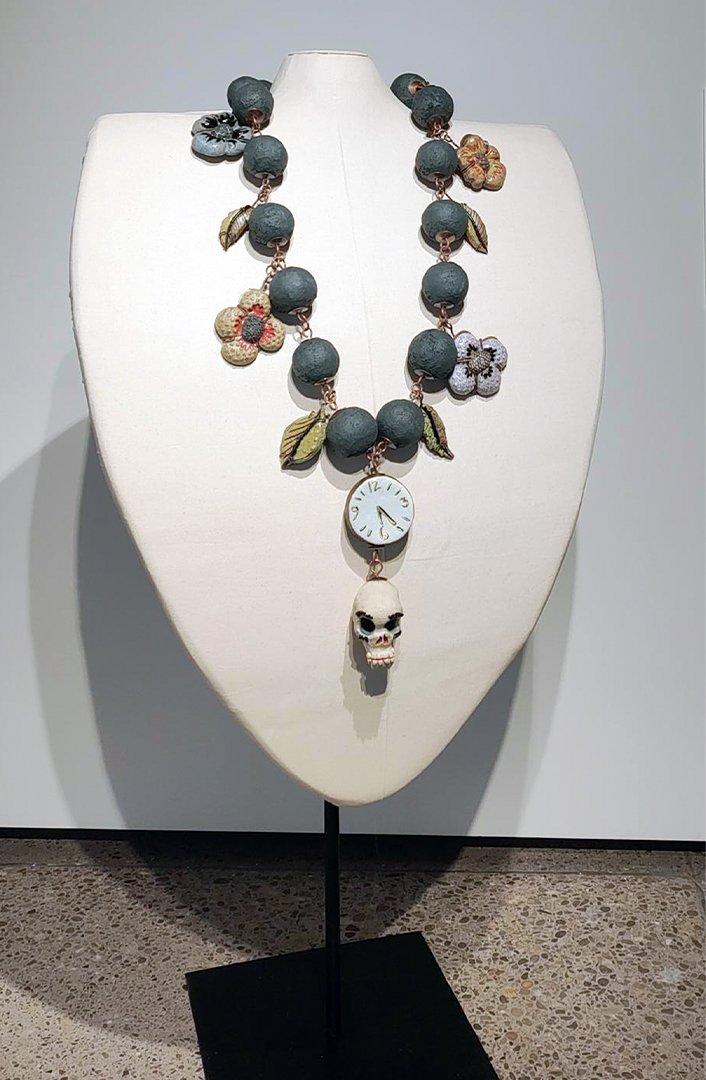 A necklace displayed on a white bust, featuring large dark beads, fabric flowers, leaves, a small clock face, and a skull pendant.