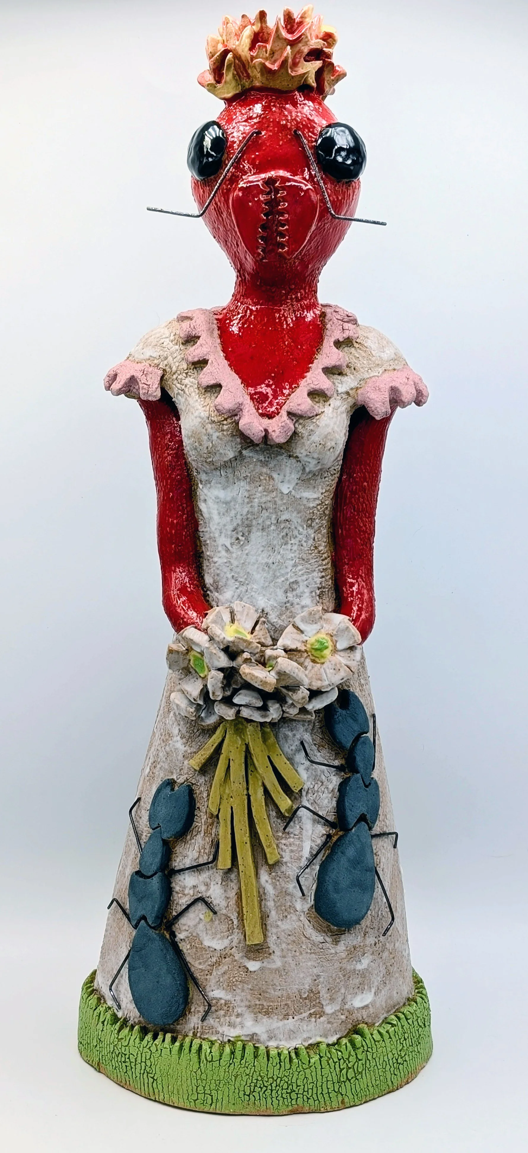 A whimsical figurine with a strawberry head, black eyes, ant-like legs, and a dress with ants climbing on it, standing on a textured green base.