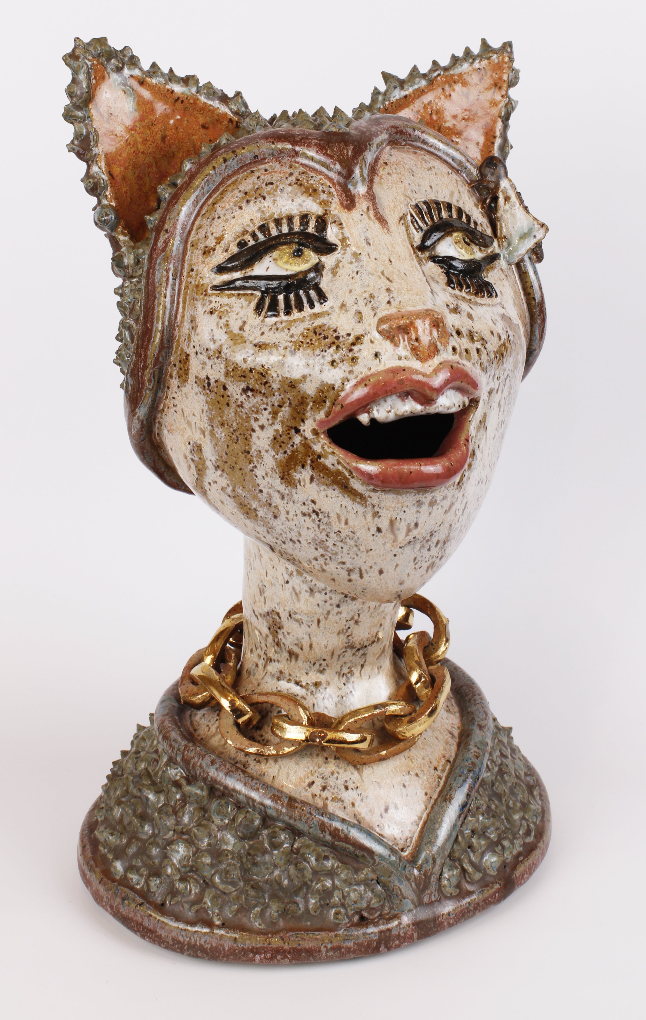 Ceramic sculpture of a woman's head with exaggerated facial features, cat ears, and a gold chain necklace, set against a plain background.