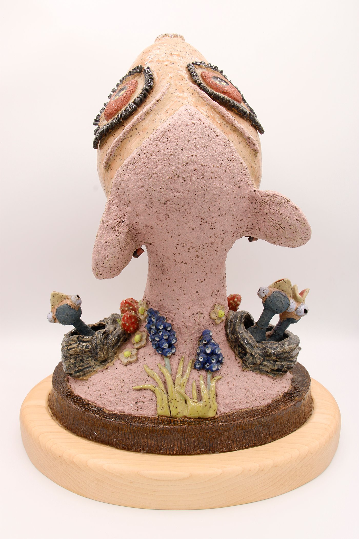 Colorful ceramic sculpture of a smiling frog with decorative flowers and mushrooms at its base, mounted on a wooden stand.