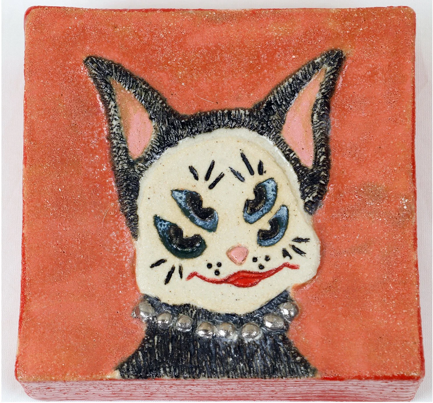 A decorative tile featuring a cartoonish black and white cat with blue eyes, a pink nose, red lips, and a necklace of silver beads, set against a reddish-orange background.